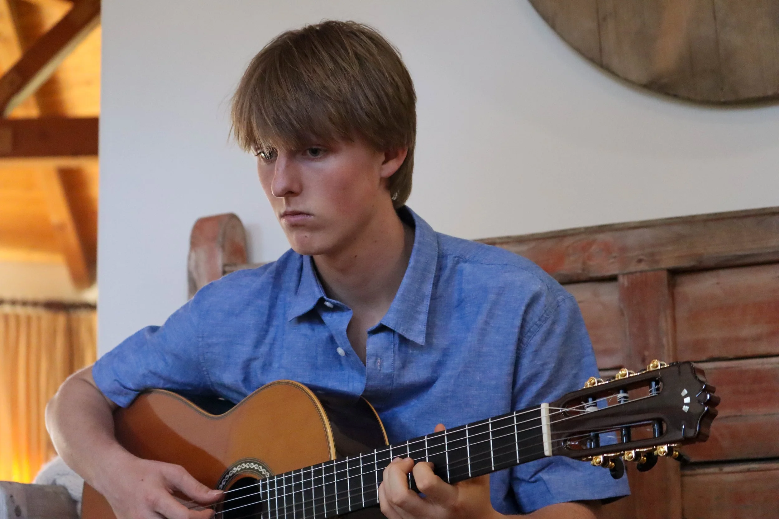 A young man with brown hair wearing a blue shirt playing an acoustic guitar indoors.