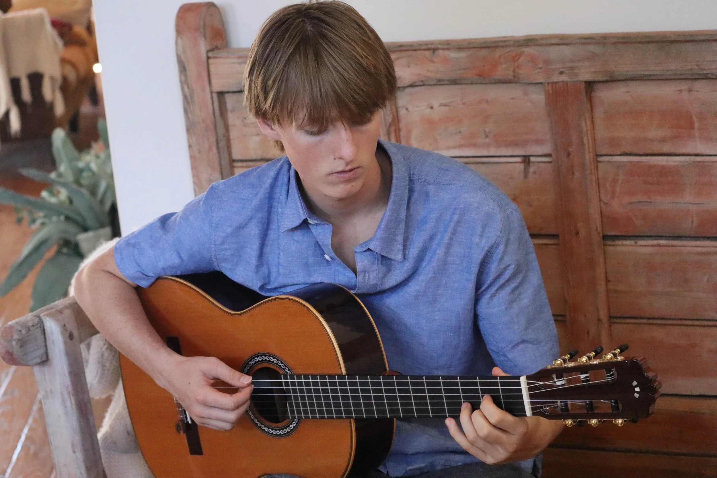 A young man with brown hair wearing a blue collared shirt is playing an acoustic guitar indoors near a wooden fence and a potted plant.
