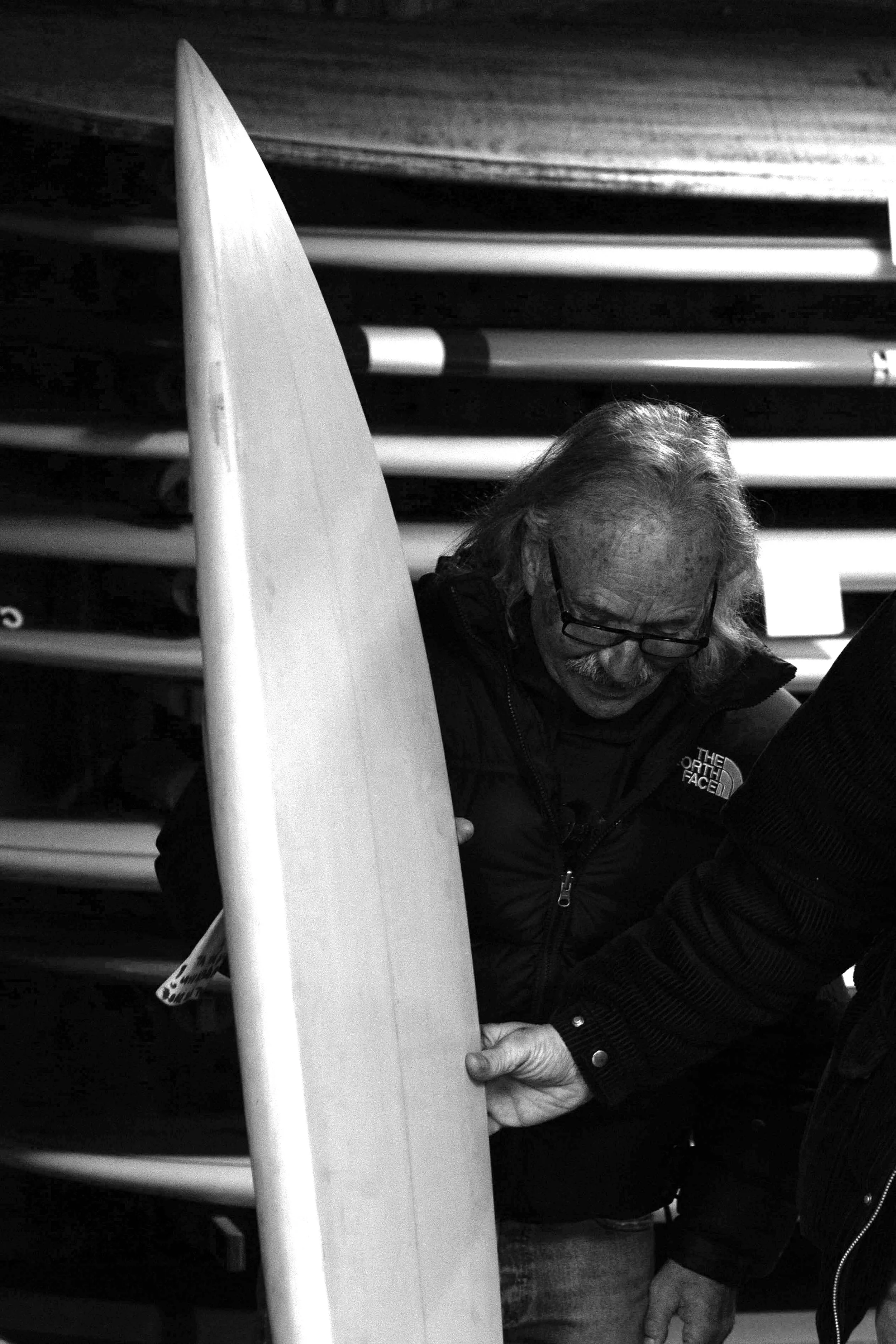 A man with glasses and long hair holding a surfboard indoors, with shelves of other surfboards in the background.