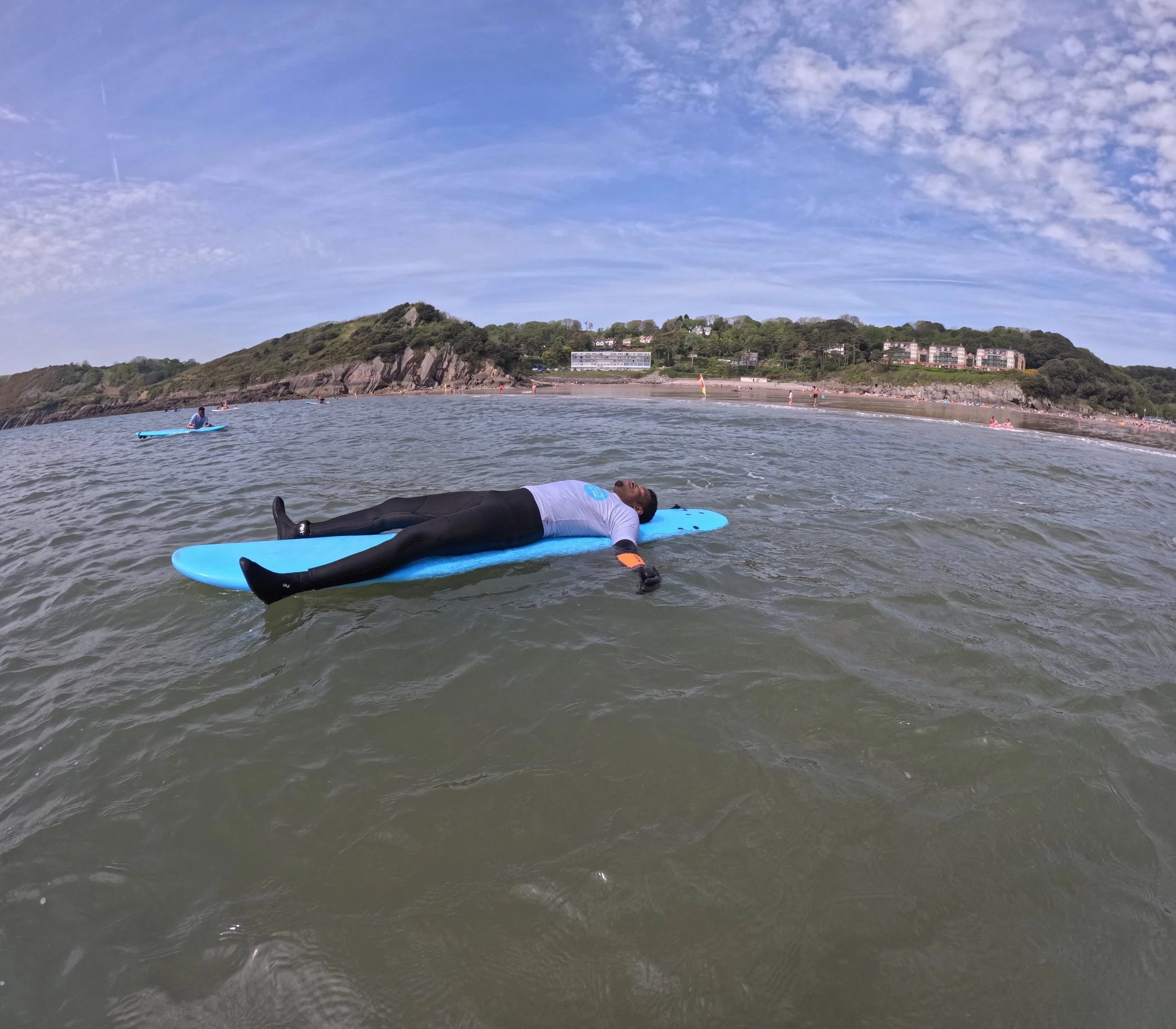Person lying flat on a blue surfboard in the water, wearing a wetsuit and gloves, with a beach and hillside in the background under a partly cloudy sky.