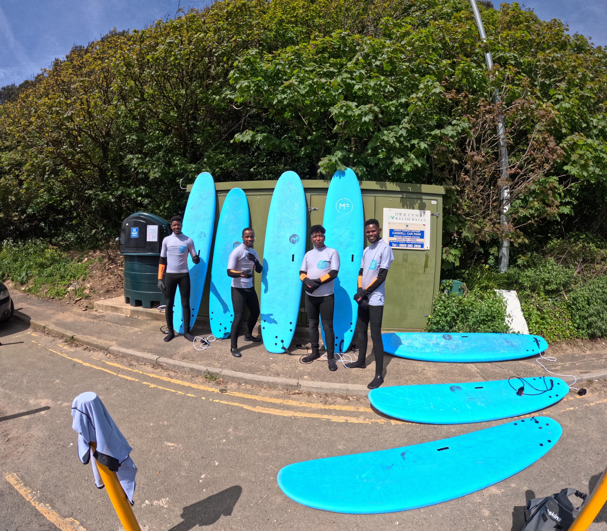 Four people dressed in surf gear standing next to five blue surfboards upright on the sidewalk, with three more surfboards lying on the ground nearby, against a background of trees and a green utility box.