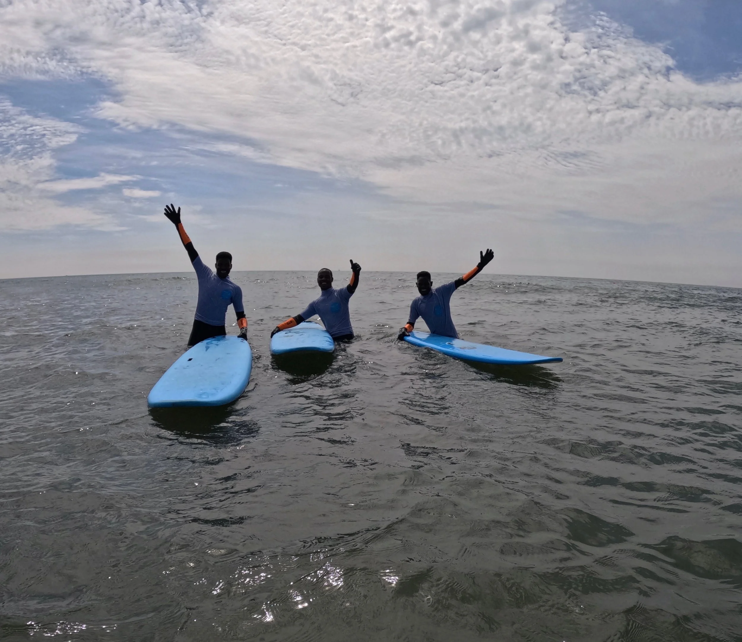 Three people in wetsuits sitting on surfboards in the ocean, waving at the camera, under a partly cloudy sky.