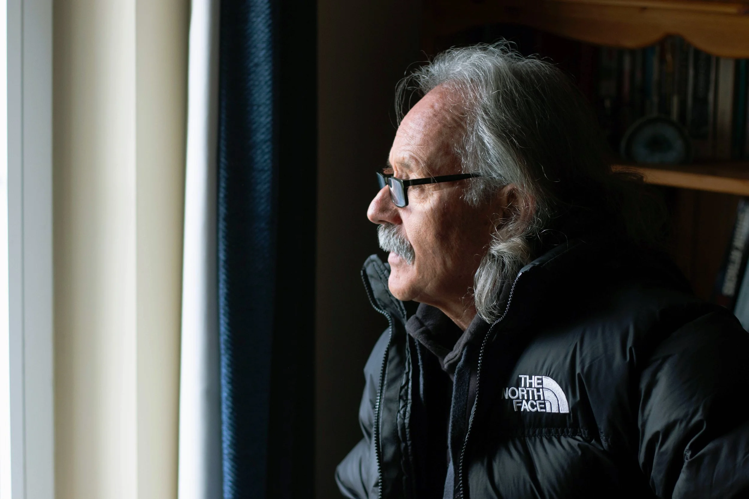 Older man with gray hair, glasses, and mustache looking out a window on a cloudy day, wearing a black North Face jacket.