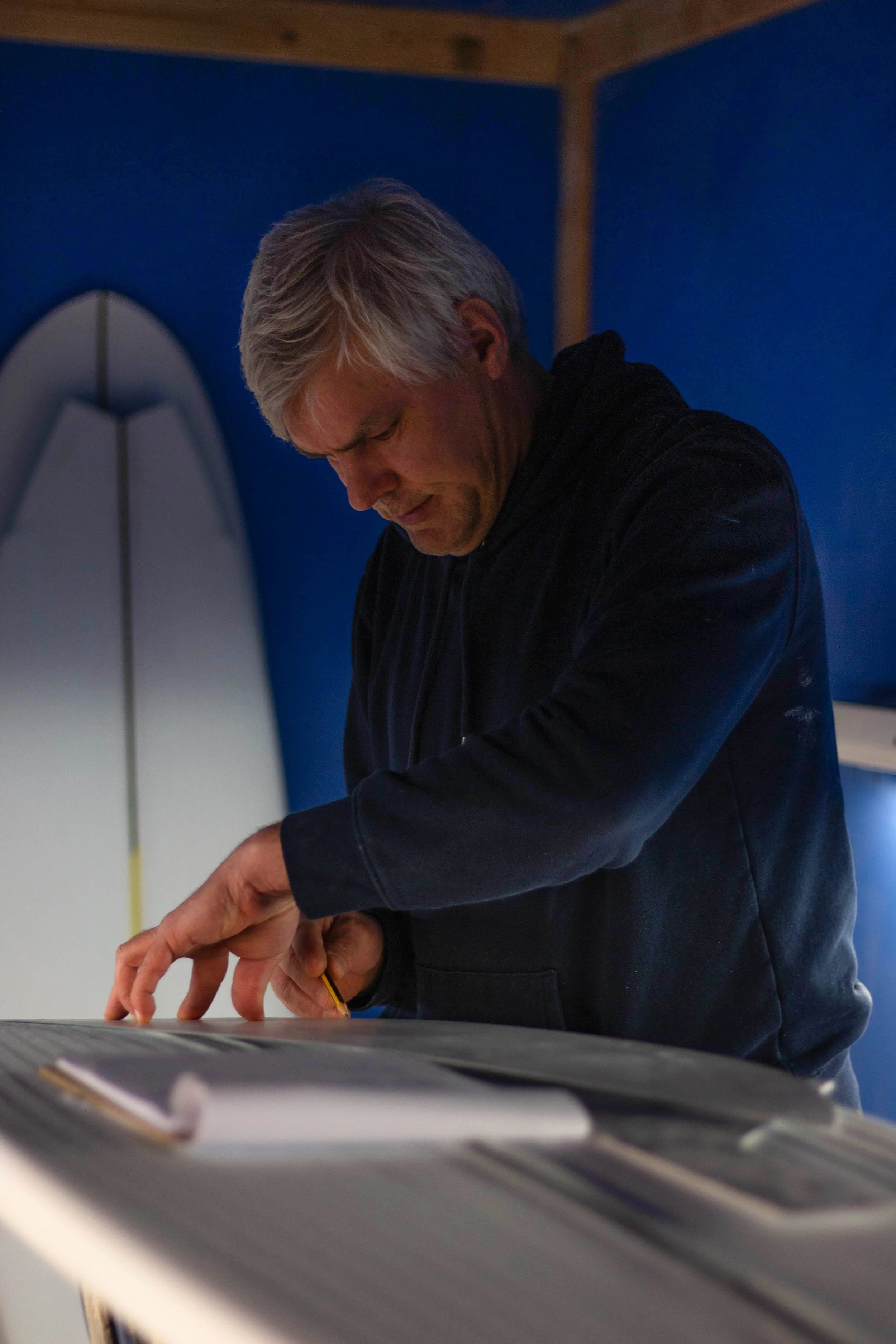 A man leans over a surfboard, meauring out and preforming the shaping process.