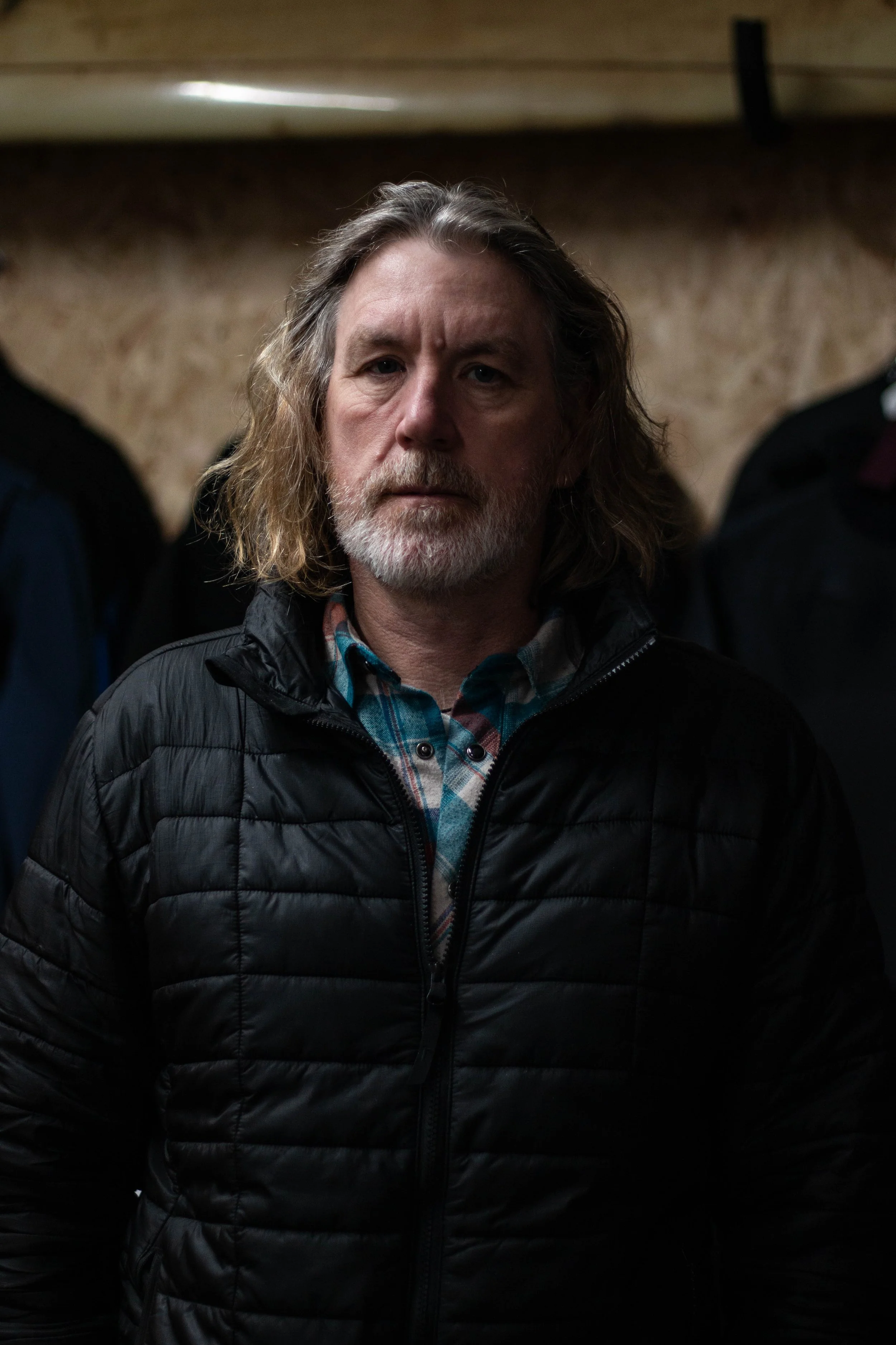 A middle-aged man with long, wavy gray hair and a beard, wearing a black quilted jacket over a plaid shirt, standing indoors with a neutral expression.