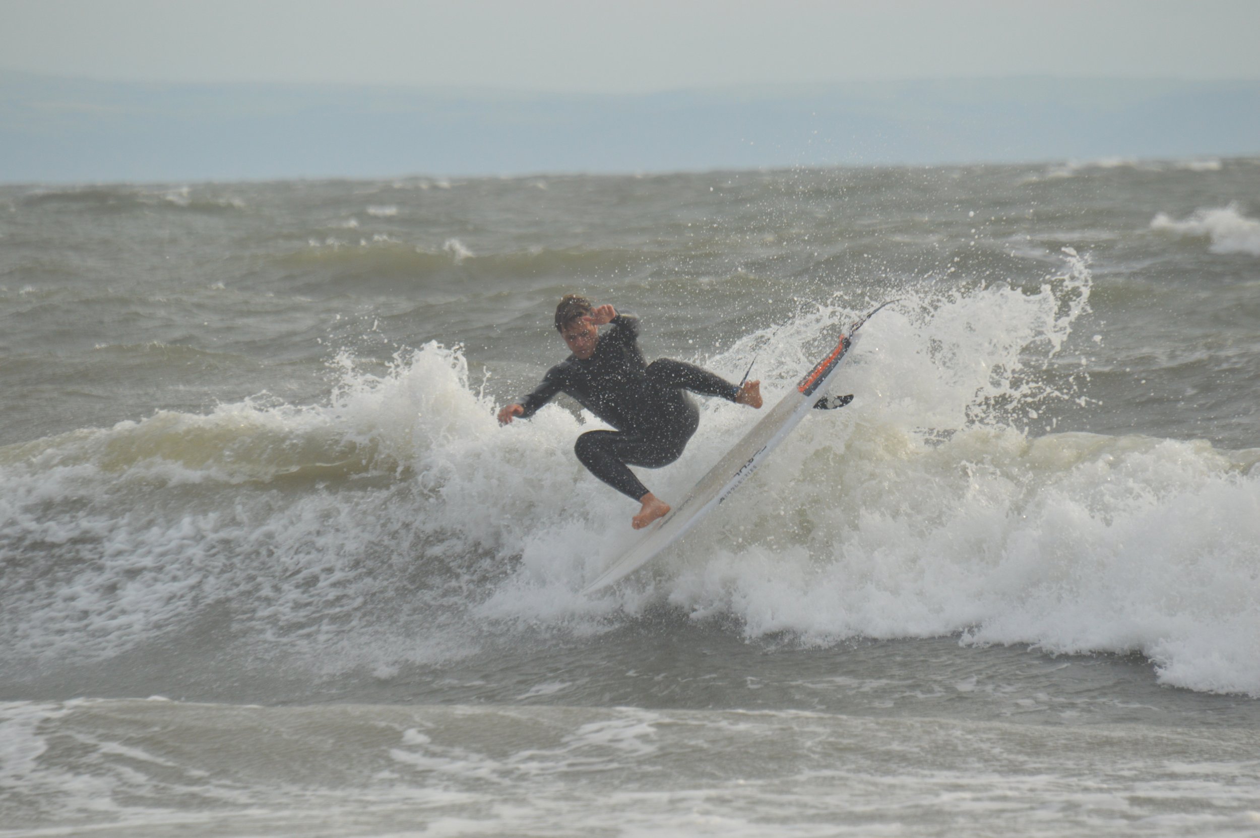 Person surfing on a wave in the ocean, wearing a black wetsuit, with a cloudy sky in the background.