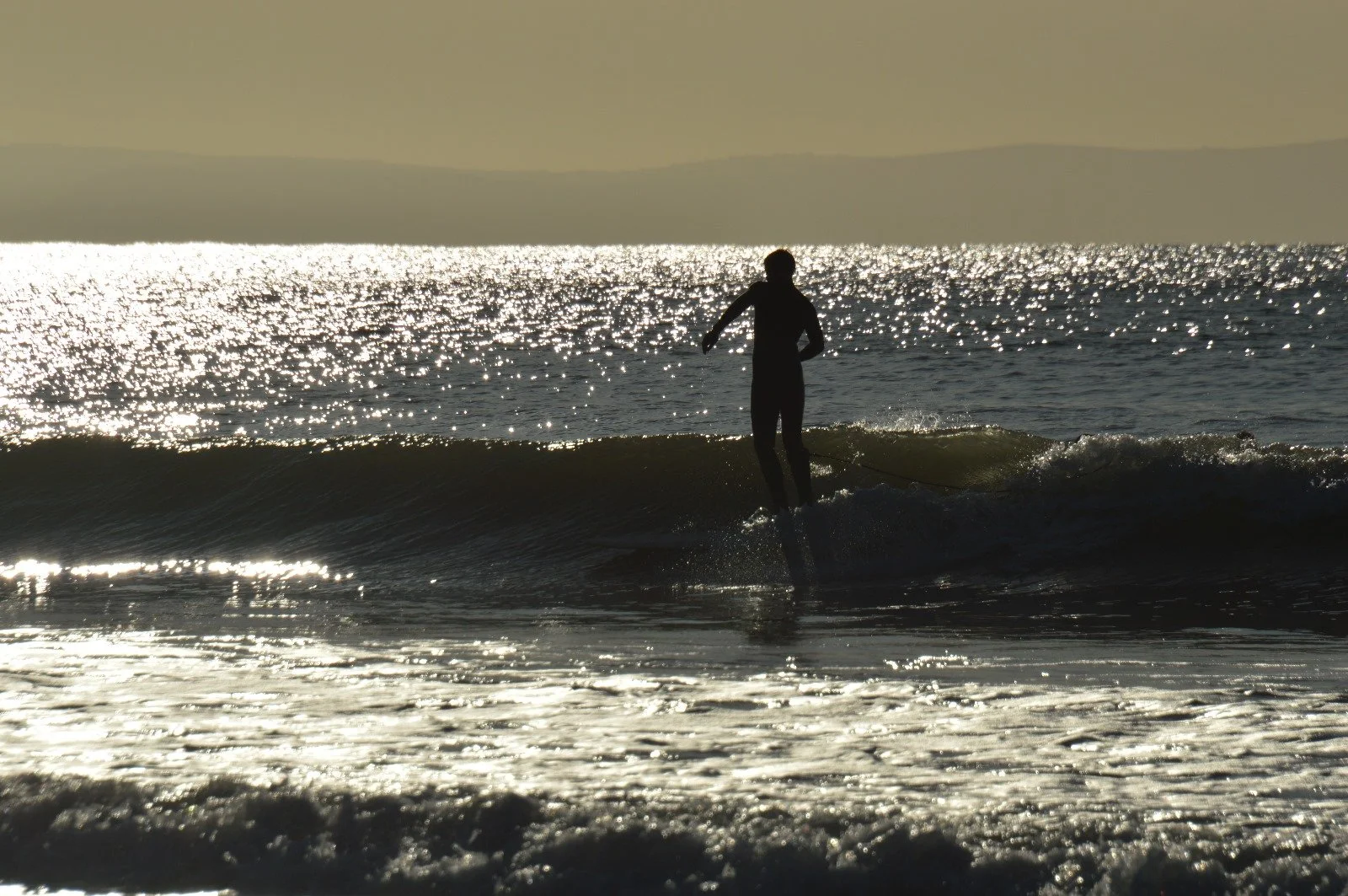 A person surfing on a wave during sunset with reflections of sunlight on the ocean.