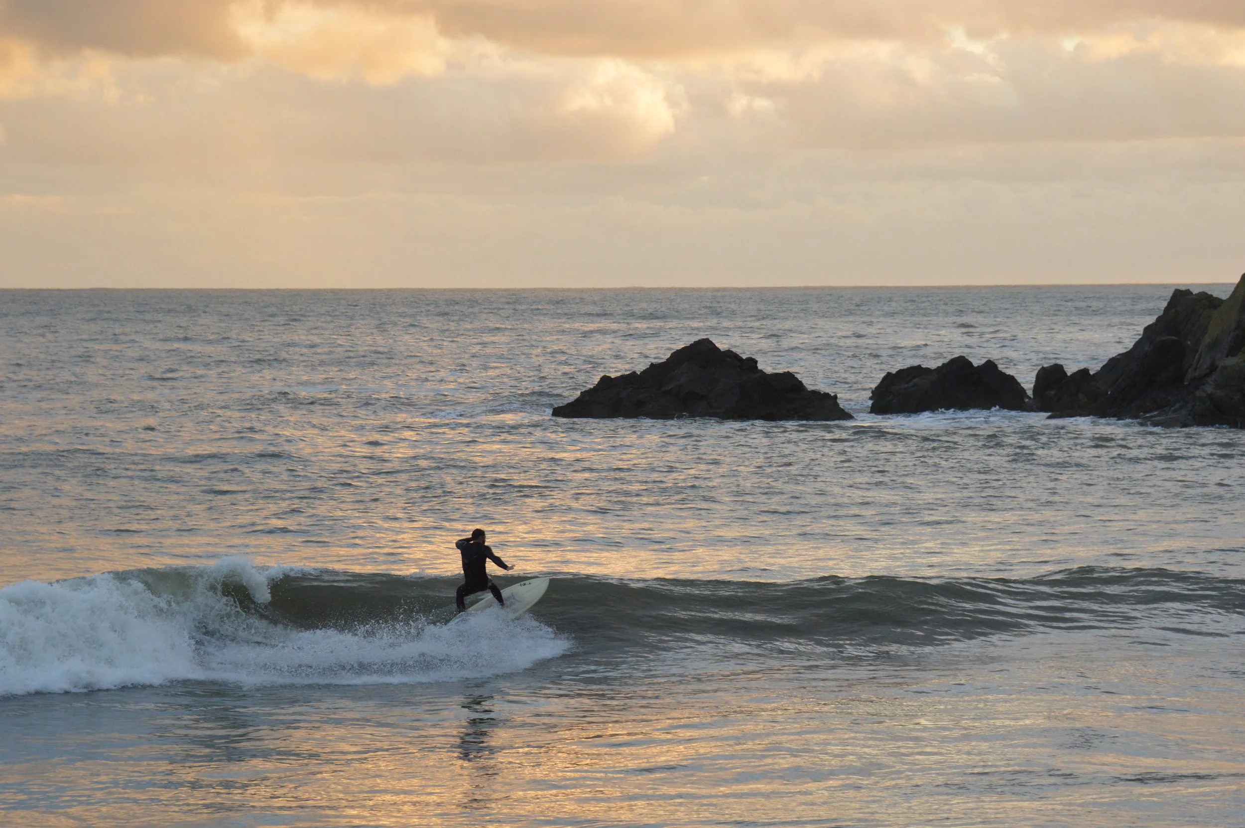 A person surfing on a wave near rocky formations in the ocean during sunset or sunrise.
