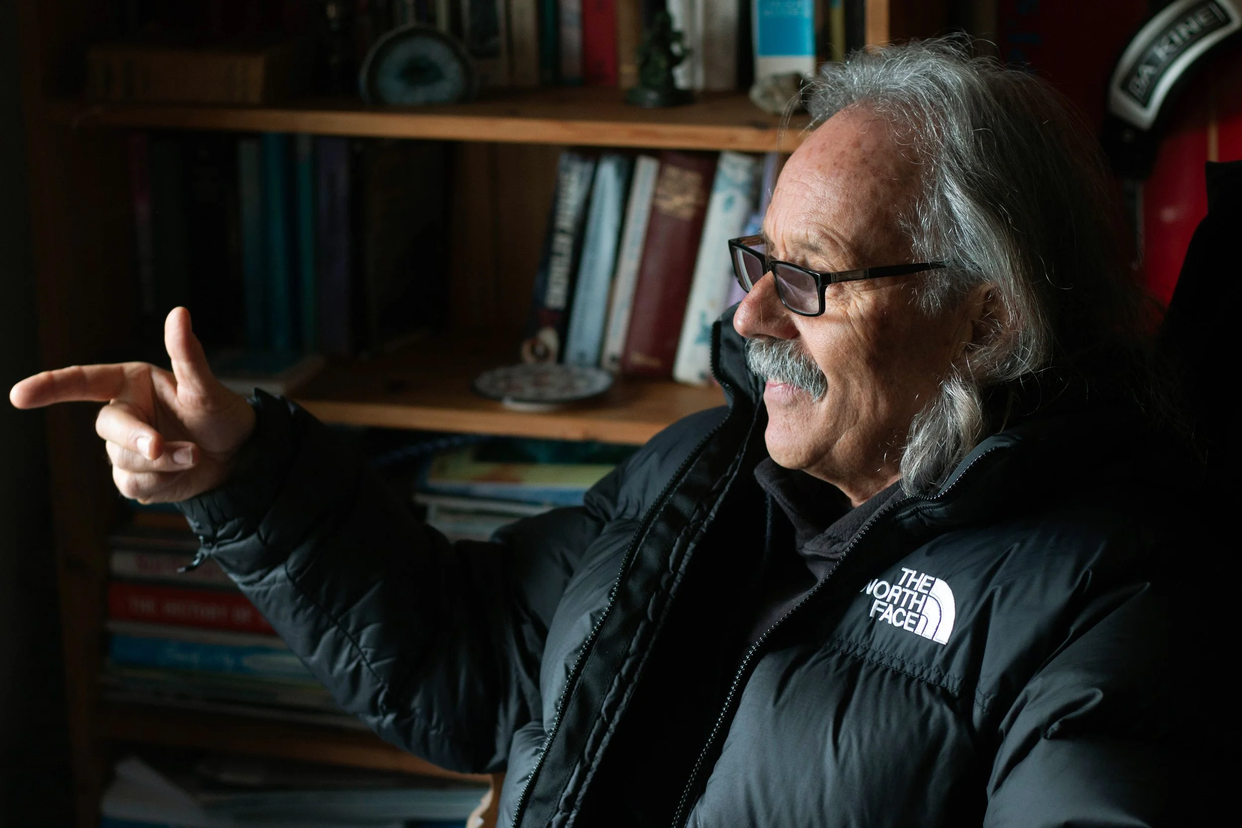 An elderly man with long gray hair and glasses, wearing a black North Face jacket, sitting in a room with bookshelves, pointing and smiling.