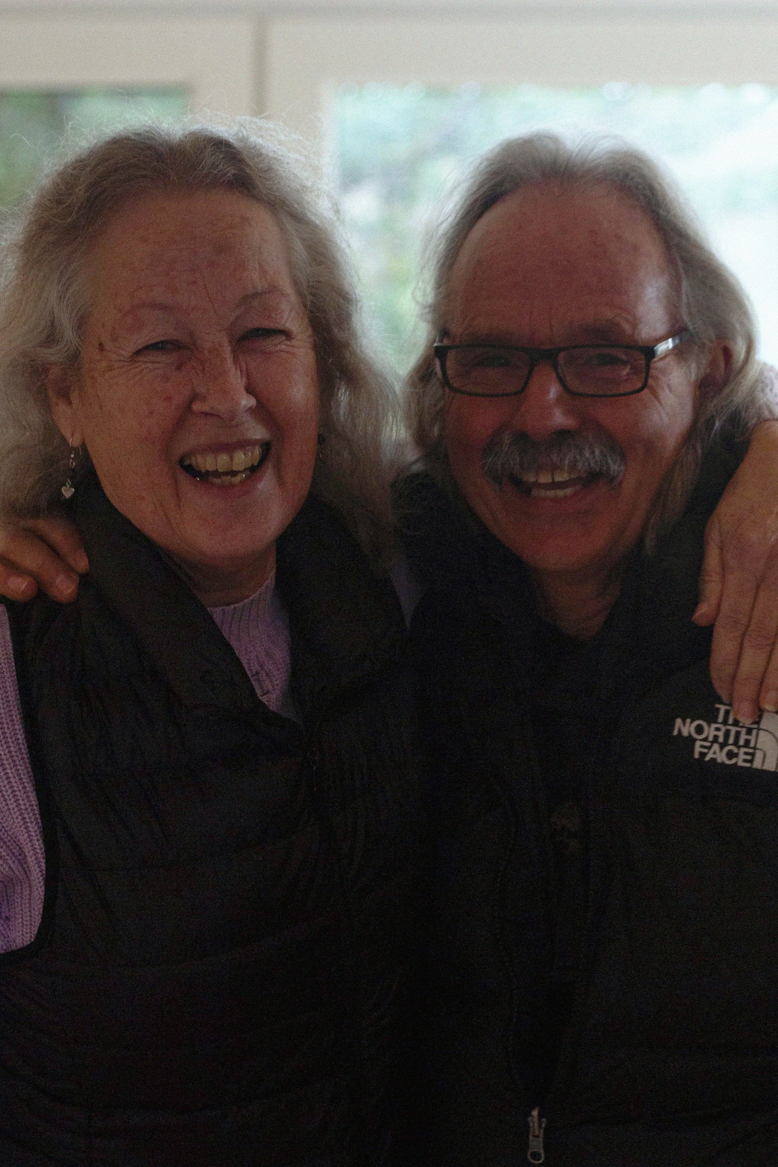 Two older adults, a woman and a man, smiling and embracing each other indoors, with a blurred window in the background.