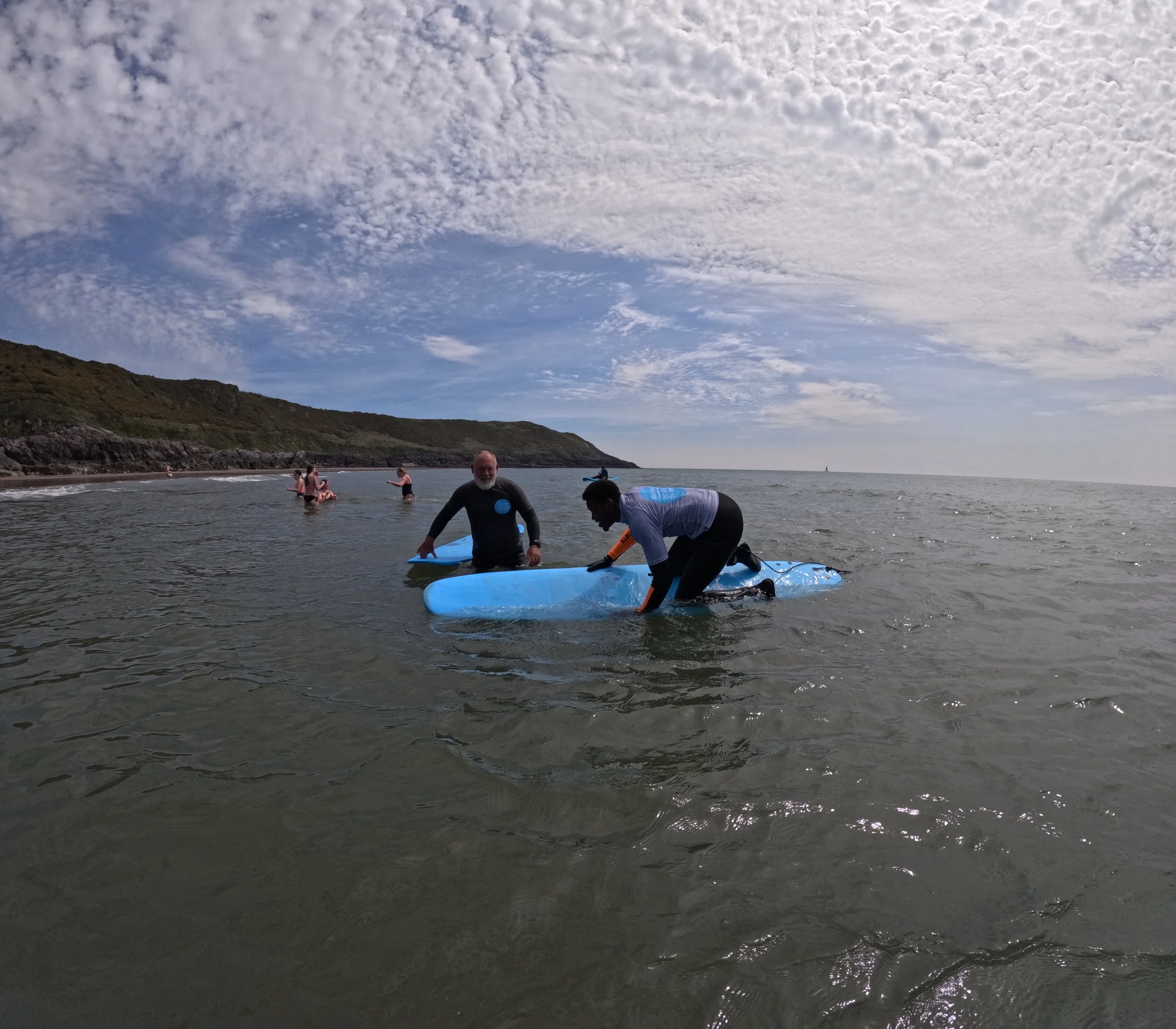 People at the beach, some standing in the water with surfboards and others sitting on the shore, under a partly cloudy sky.