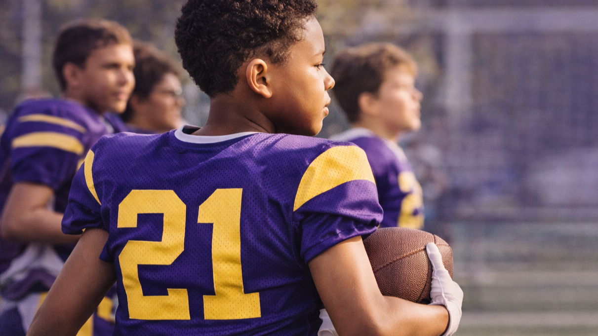 A young African American boy wearing a purple football jersey with yellow accents and the number 21, holding a football, standing on a football field with a group of teammates in similar uniforms in the background.