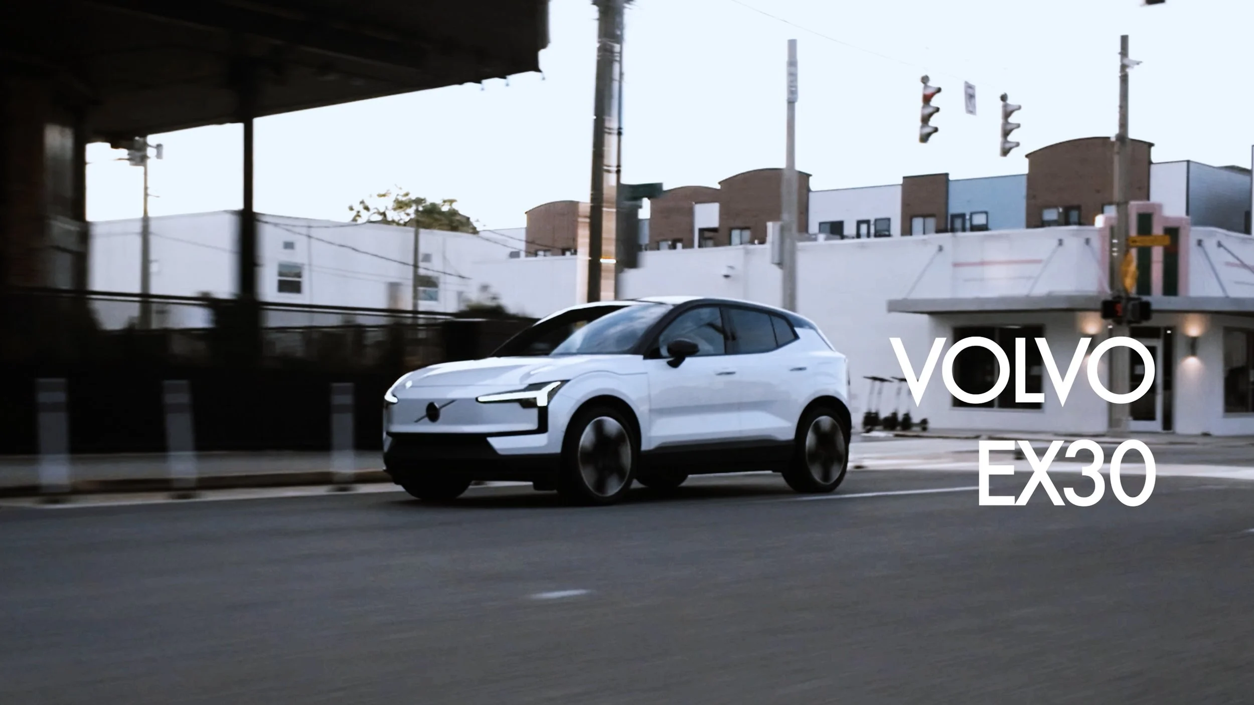 A white Volvo EX30 electric SUV driving on an urban street with modern buildings in the background.