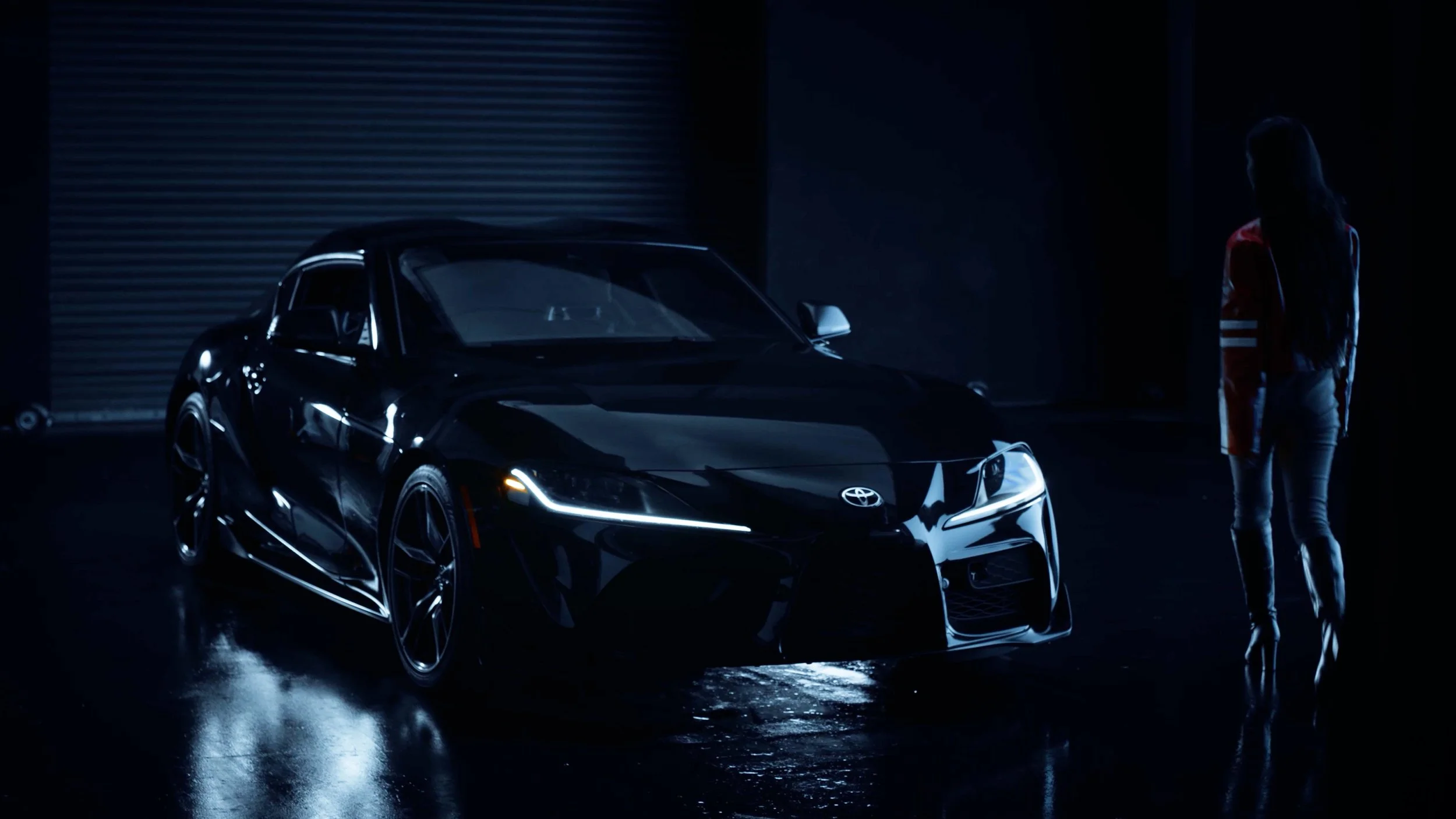 A sleek black Toyota sports car with bright headlights parked inside a dark, industrial garage with a woman standing nearby.