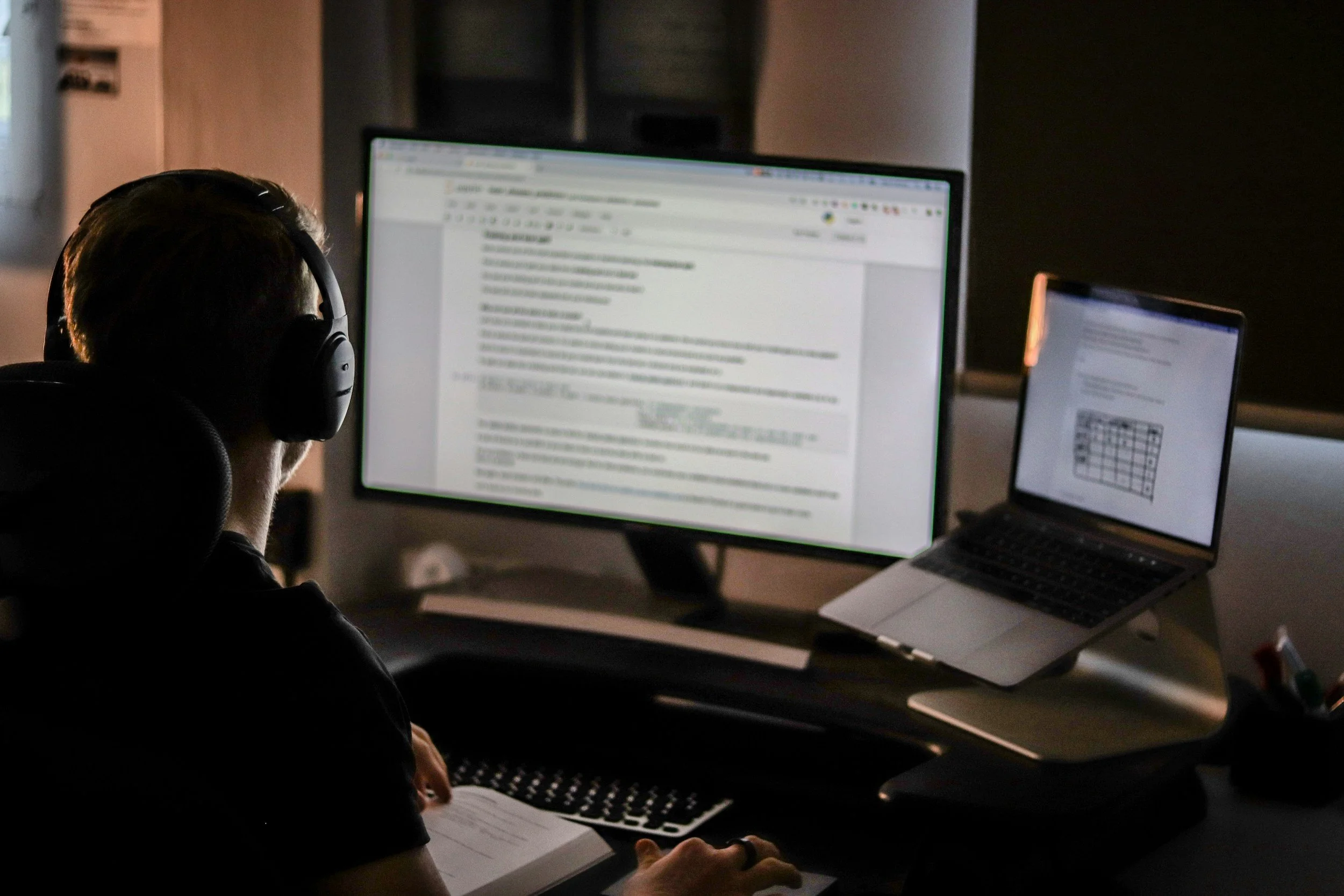 Person working at a desk with a large computer monitor, a laptop on a stand, and some papers, in a dimly lit room.
