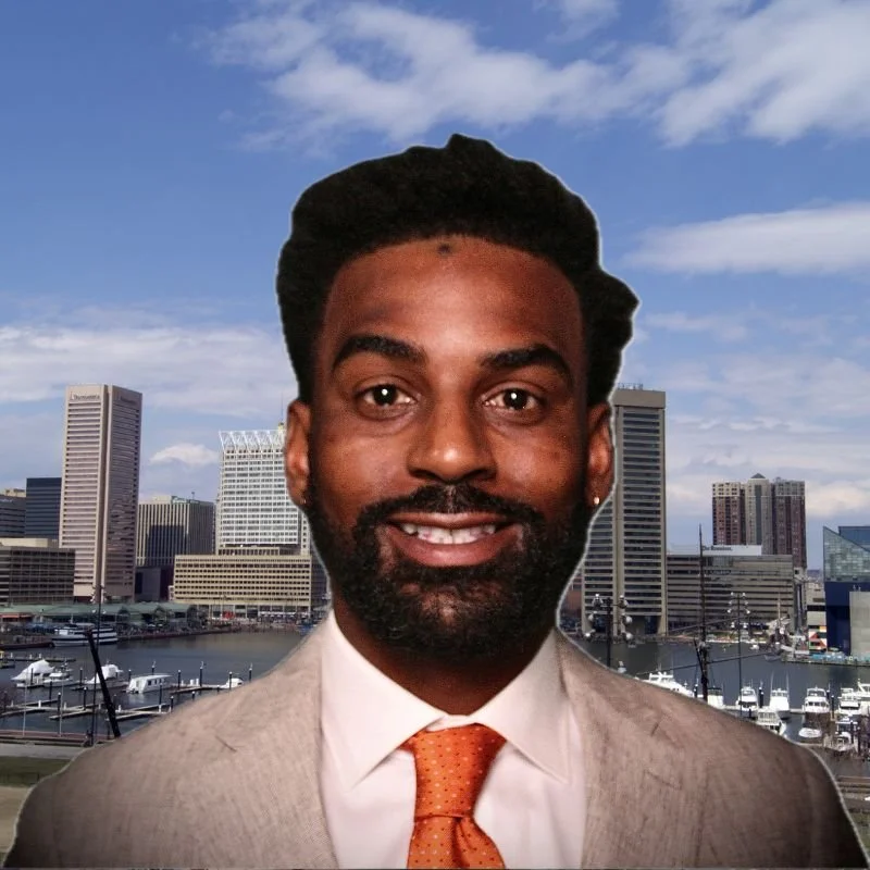 A smiling man with dark hair and beard wearing a suit and an orange tie, photo with a city skyline and harbor in the background.