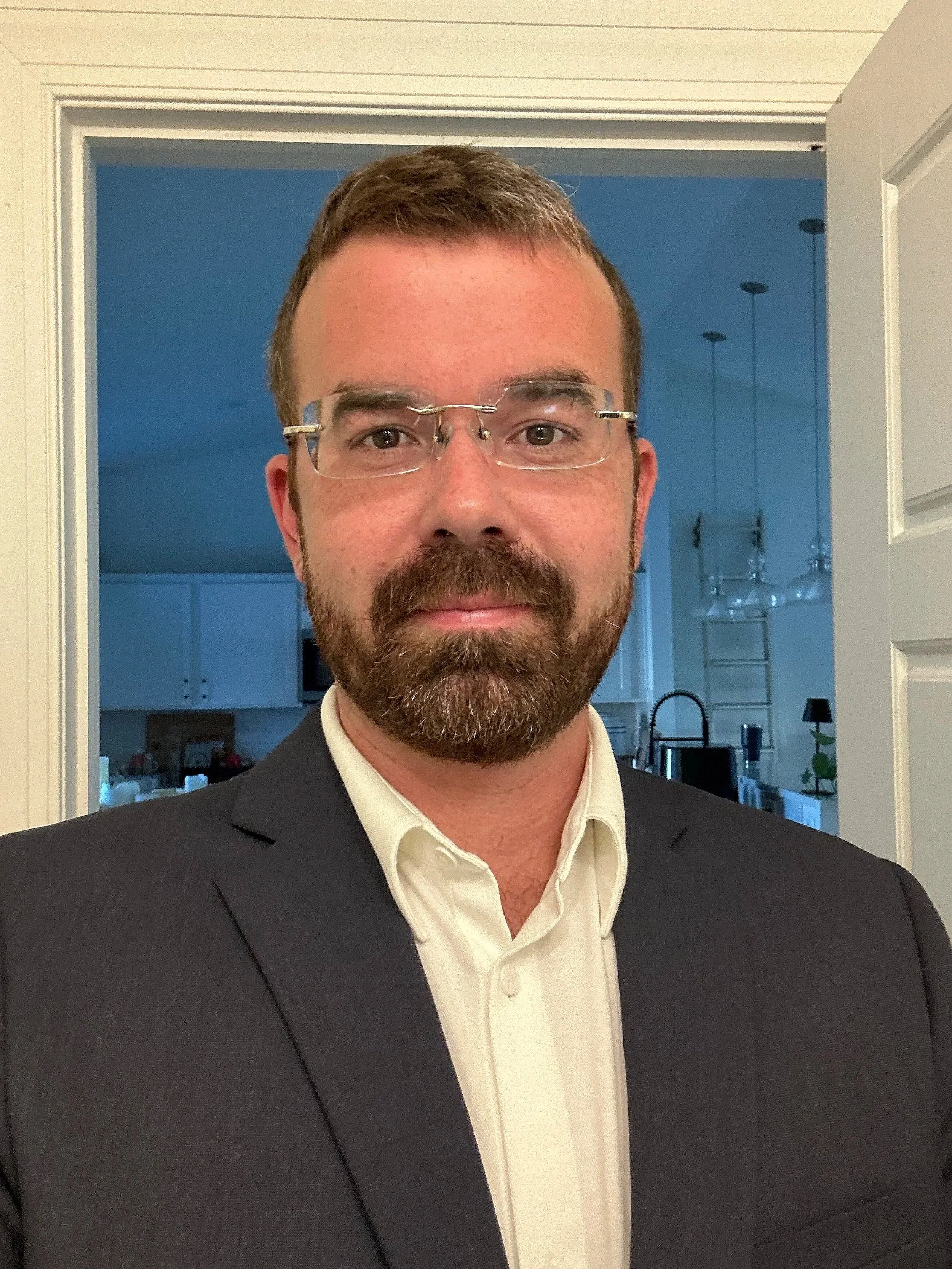 A man with glasses and a beard wearing a suit jacket and white shirt standing indoors in front of an open door, with a kitchen visible in the background.