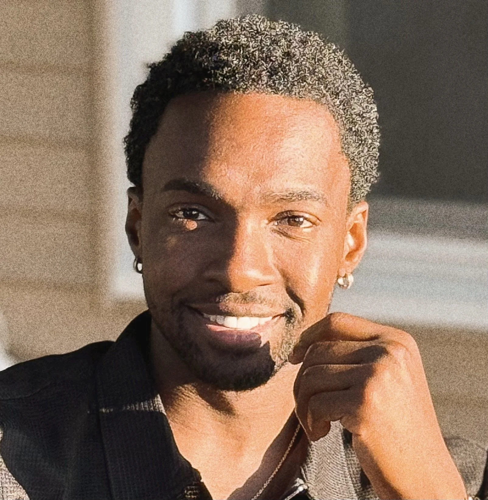 Close-up of a smiling young man with short curly hair, earrings, and a goatee, resting his chin on his hand, in natural sunlight.
