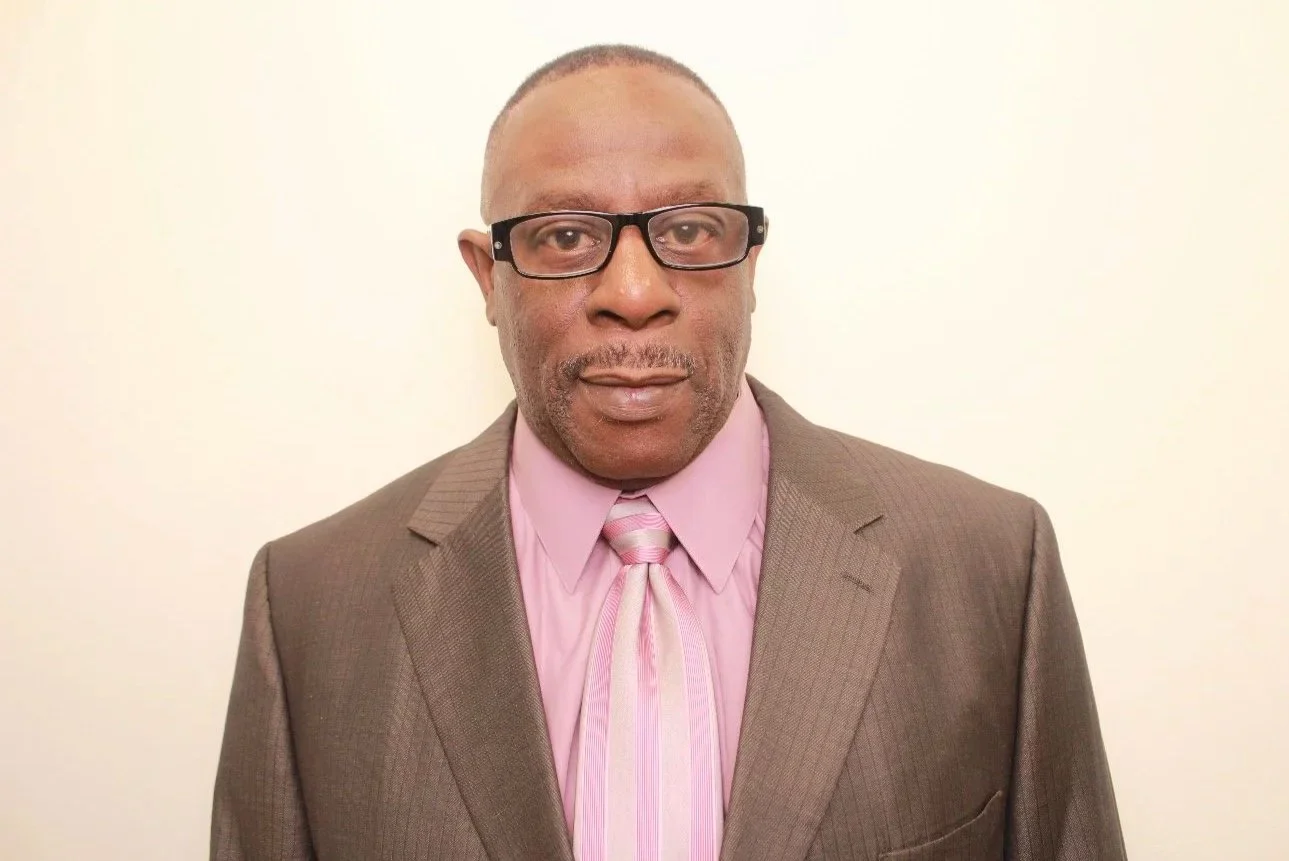 A close-up of a man with dark skin wearing sunglasses and a blue zip-up jacket, on a boat with water and trees in the background.
