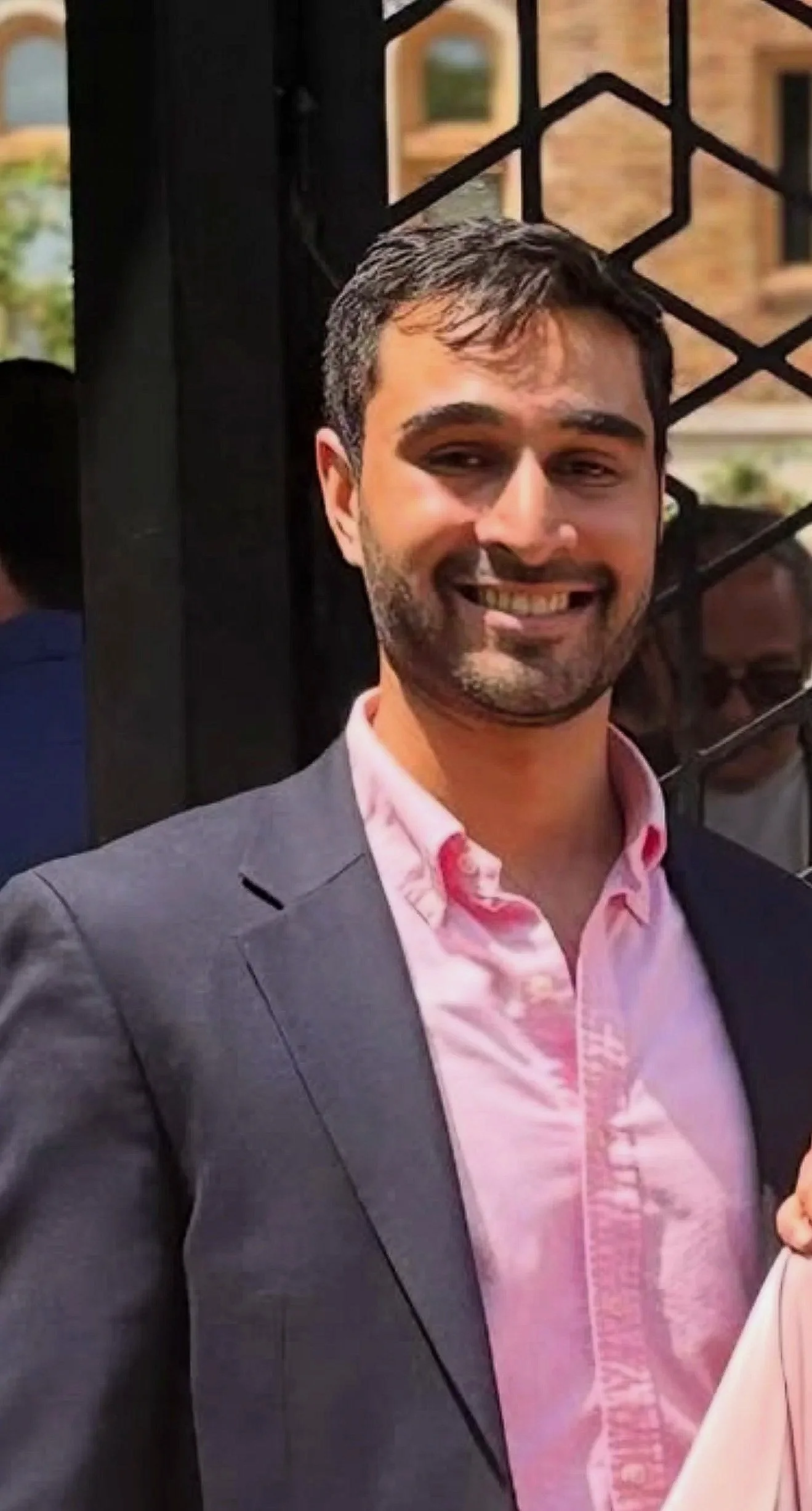 Smiling man in a gray suit and pink shirt outdoors with a decorative black iron fence and brick building in the background.