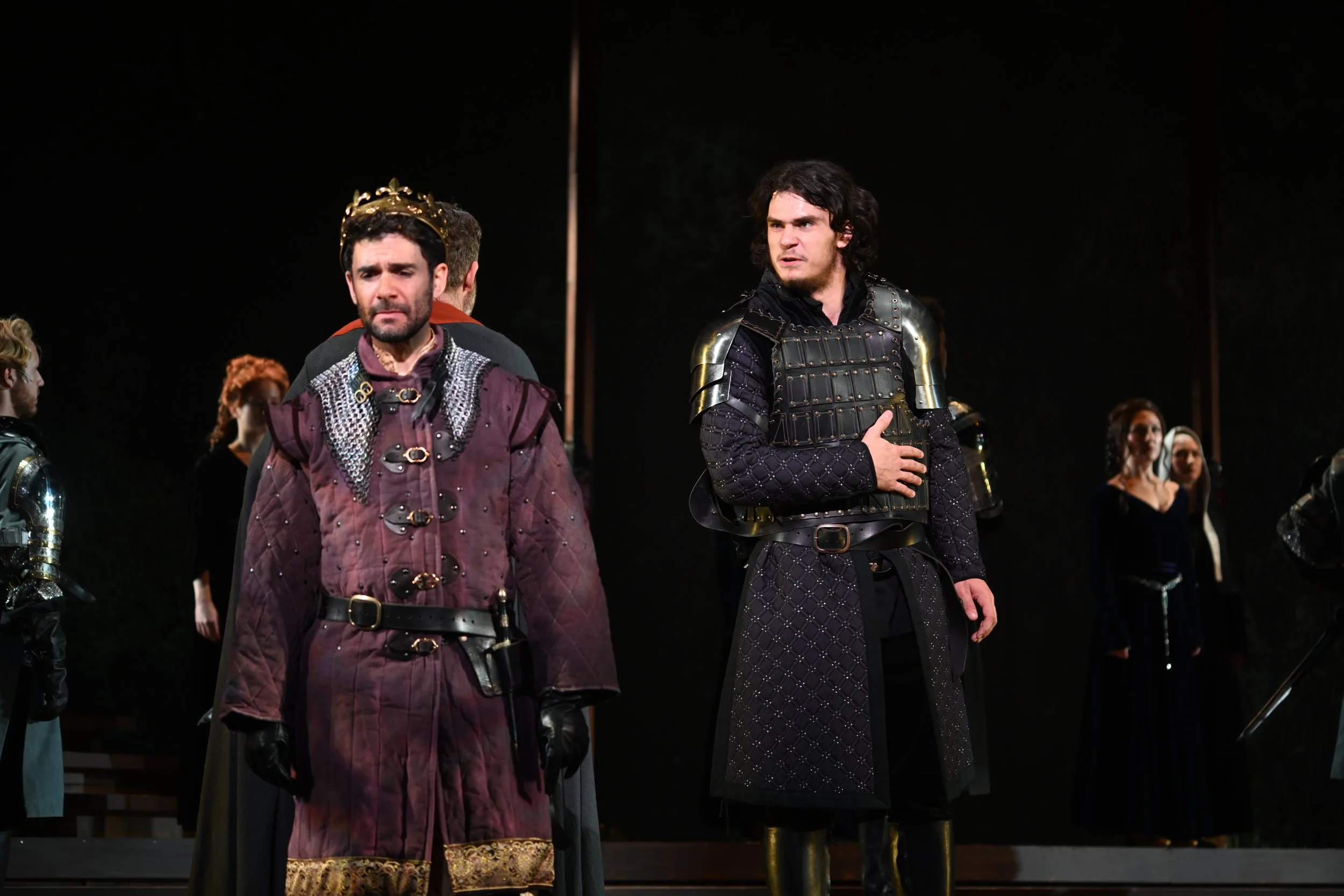 Two men dressed in medieval costume standing on a stage during a theatrical performance, with several other actors in similar costumes in the background.
