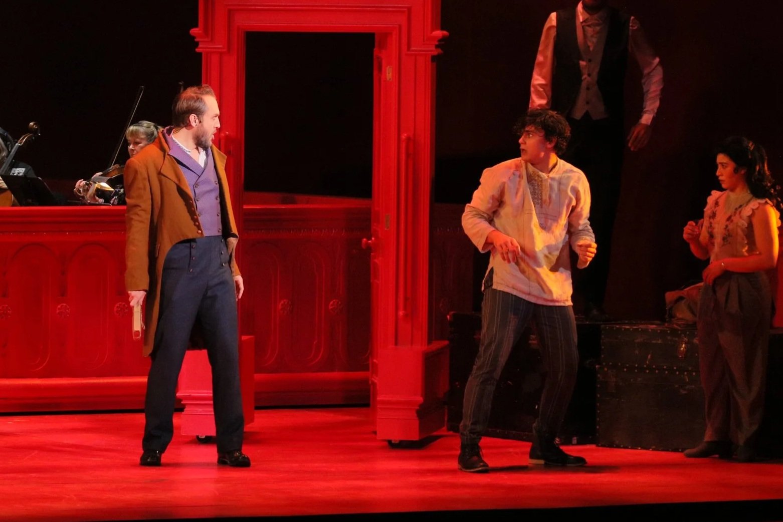 The scene shows a theatrical stage with five performers. Two men are engaged in a confrontation; one with a beard and dressed in a brown coat and blue waistcoat, holding a book, and the other with glasses and curly hair, wearing a light-colored shirt