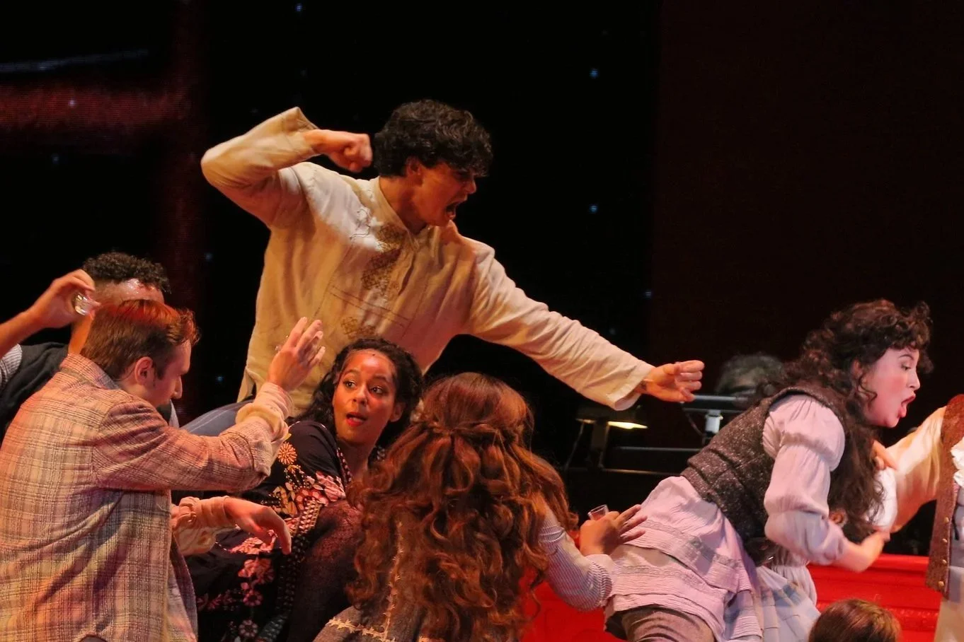 A dramatic scene from a theatrical performance with actors in period costumes, with one actor passionately shouting and others reacting intensely.