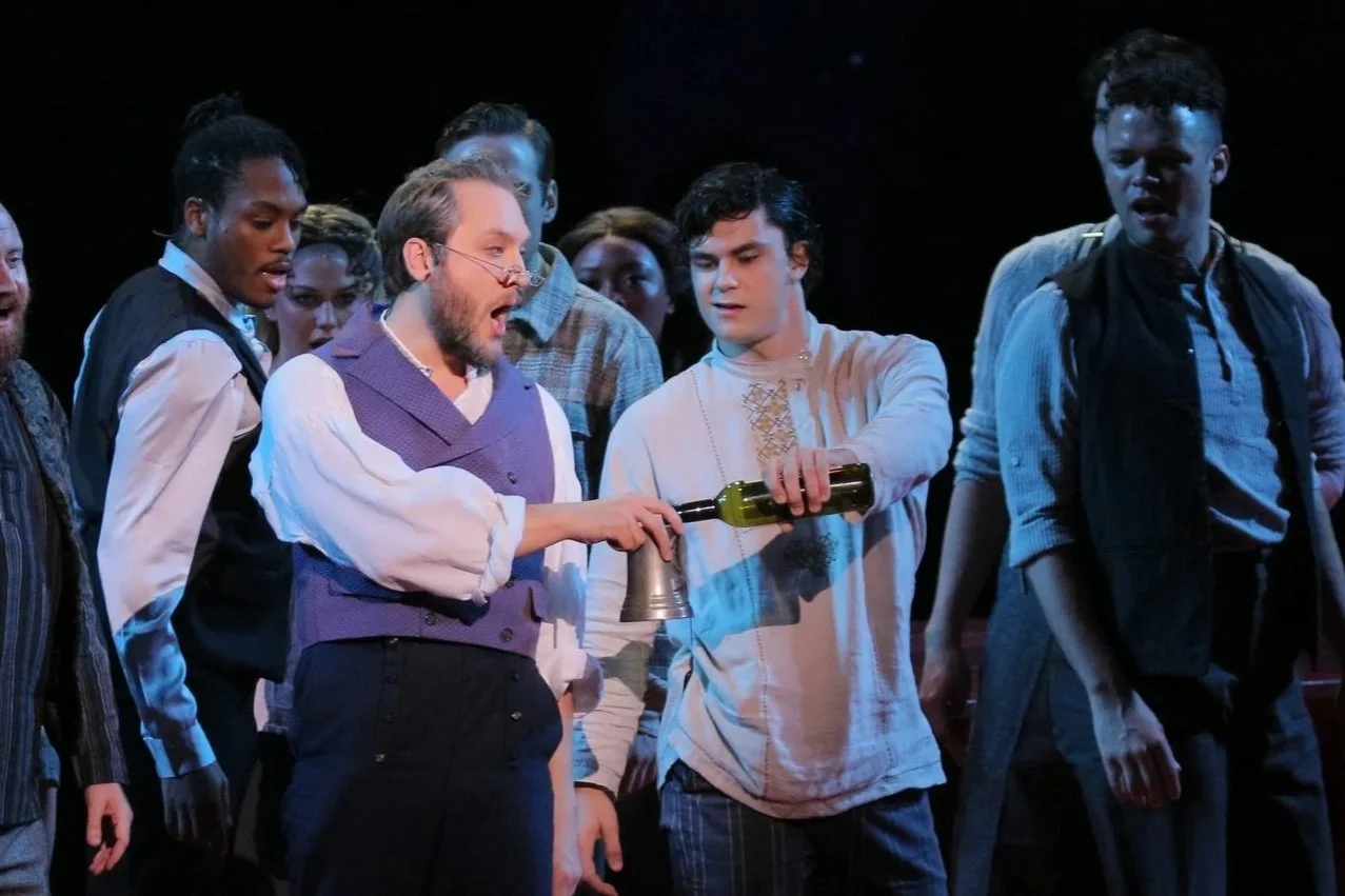 Theaters actors dressed in period costumes performing on stage, with one actor pouring what appears to be wine into a cup held by another actor.