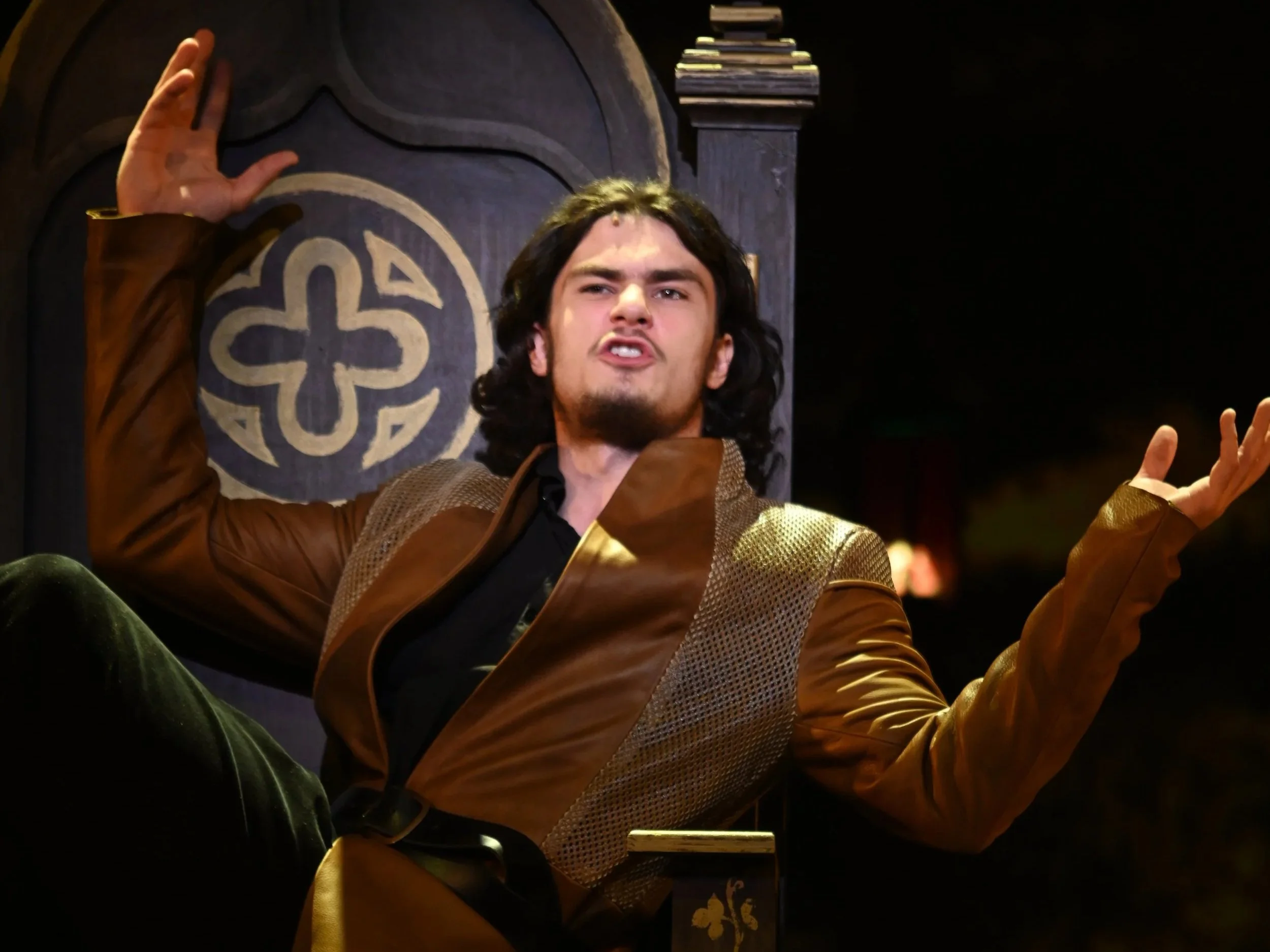 A man with long dark hair and a beard, dressed in a brown and gold medieval-style costume, is sitting on a throne-like chair and gesturing expressively with his hands.