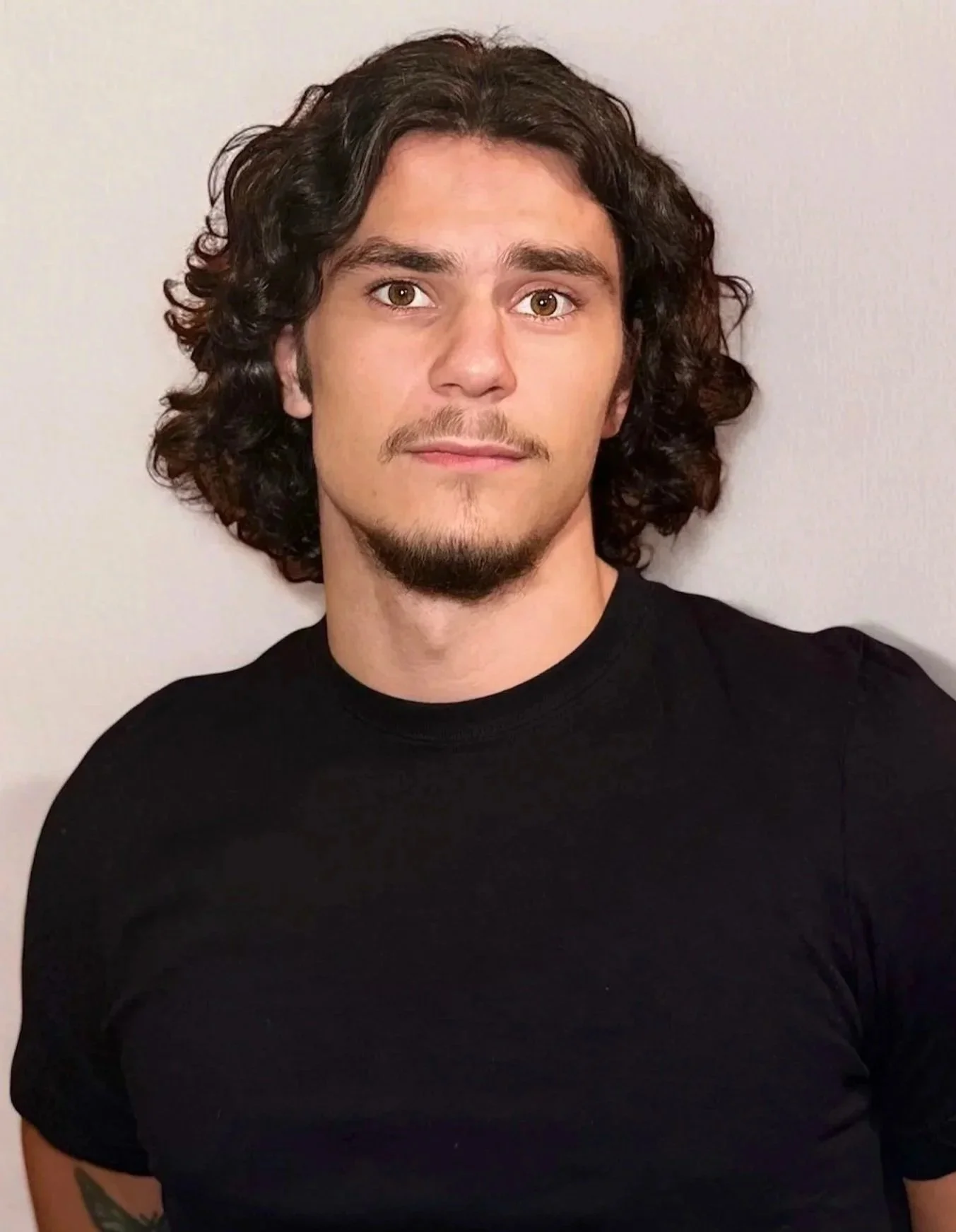 A young man with curly dark hair, light skin, and a tiny goatee wearing a black t-shirt, standing against a light-colored wall.
