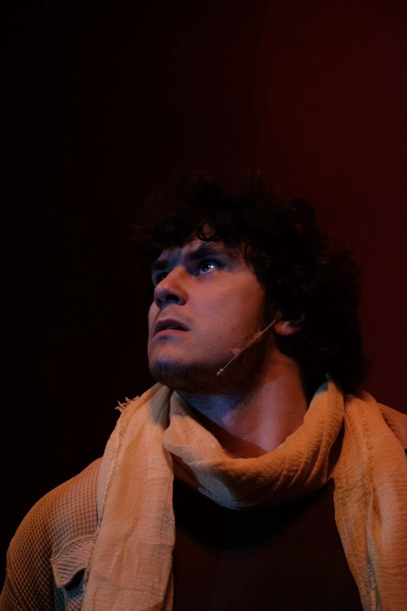 A person with dark curly hair, blue eyes, and light skin looking upward with a serious expression, wearing a beige scarf and a brown shirt, against a dark background with red lighting.