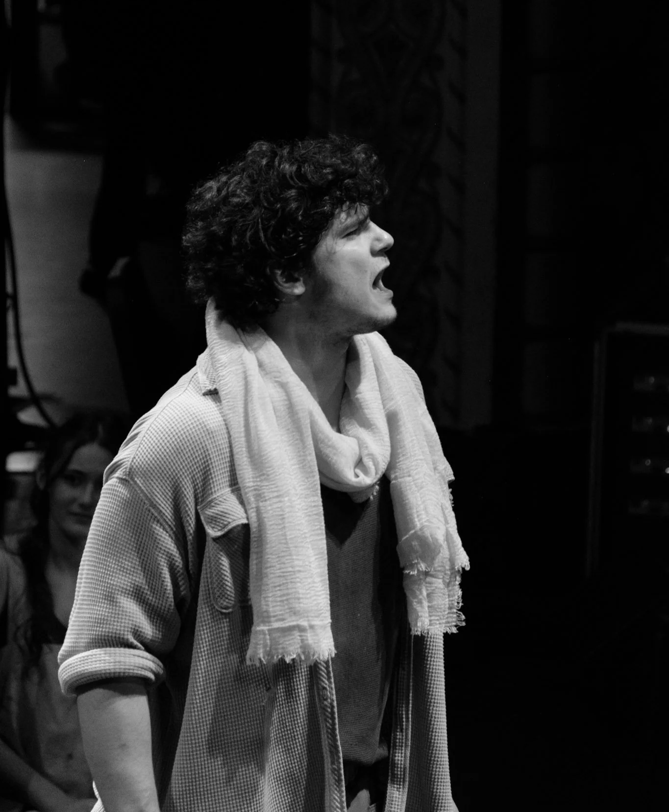 A man with curly hair singing passionately, wearing a light-colored scarf and casual clothing, in a dark room with a woman sitting in the background.