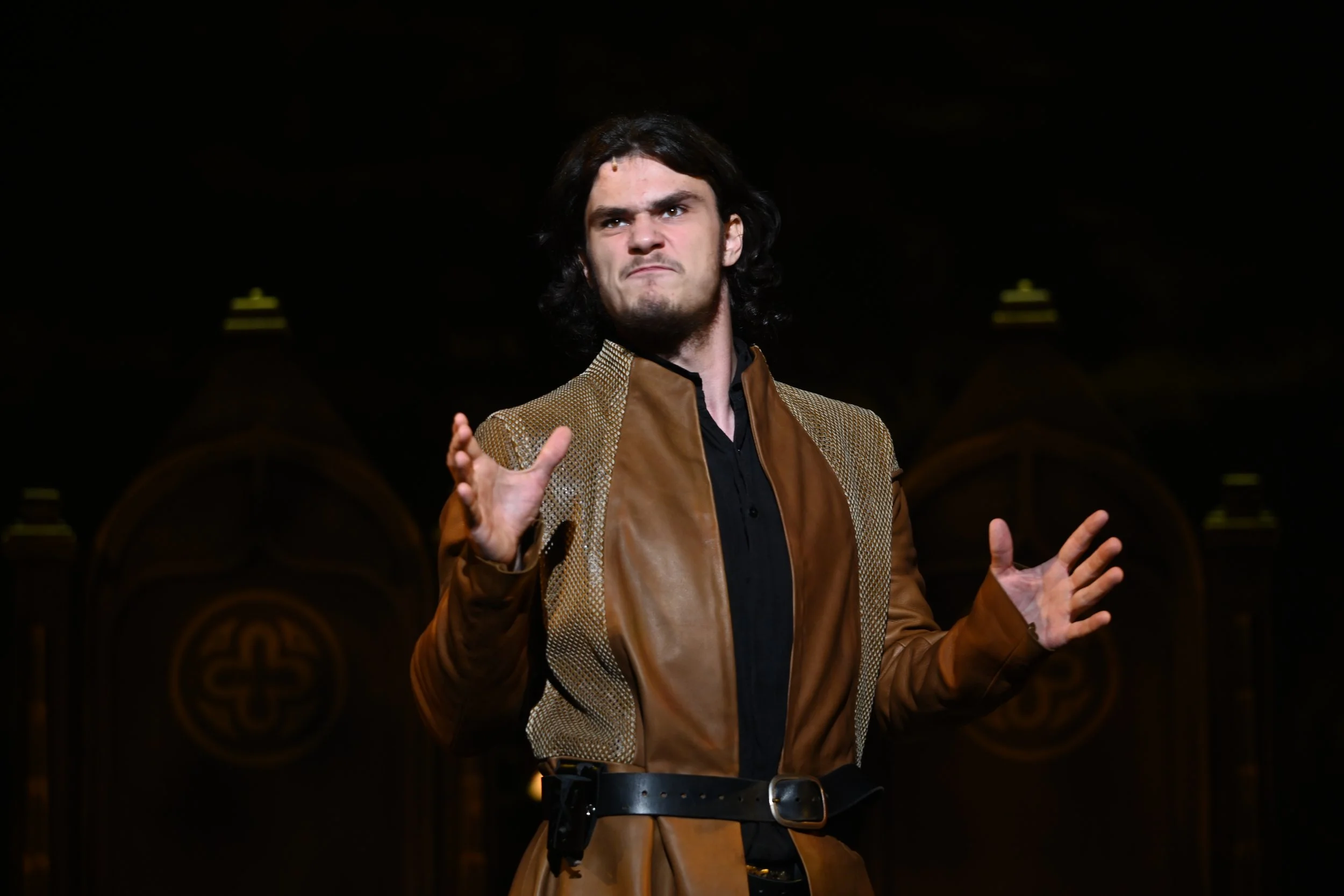 A man with dark hair and a beard making an expressive face, wearing a brown and gold jacket and standing in front of a dark background with ornate gold details.