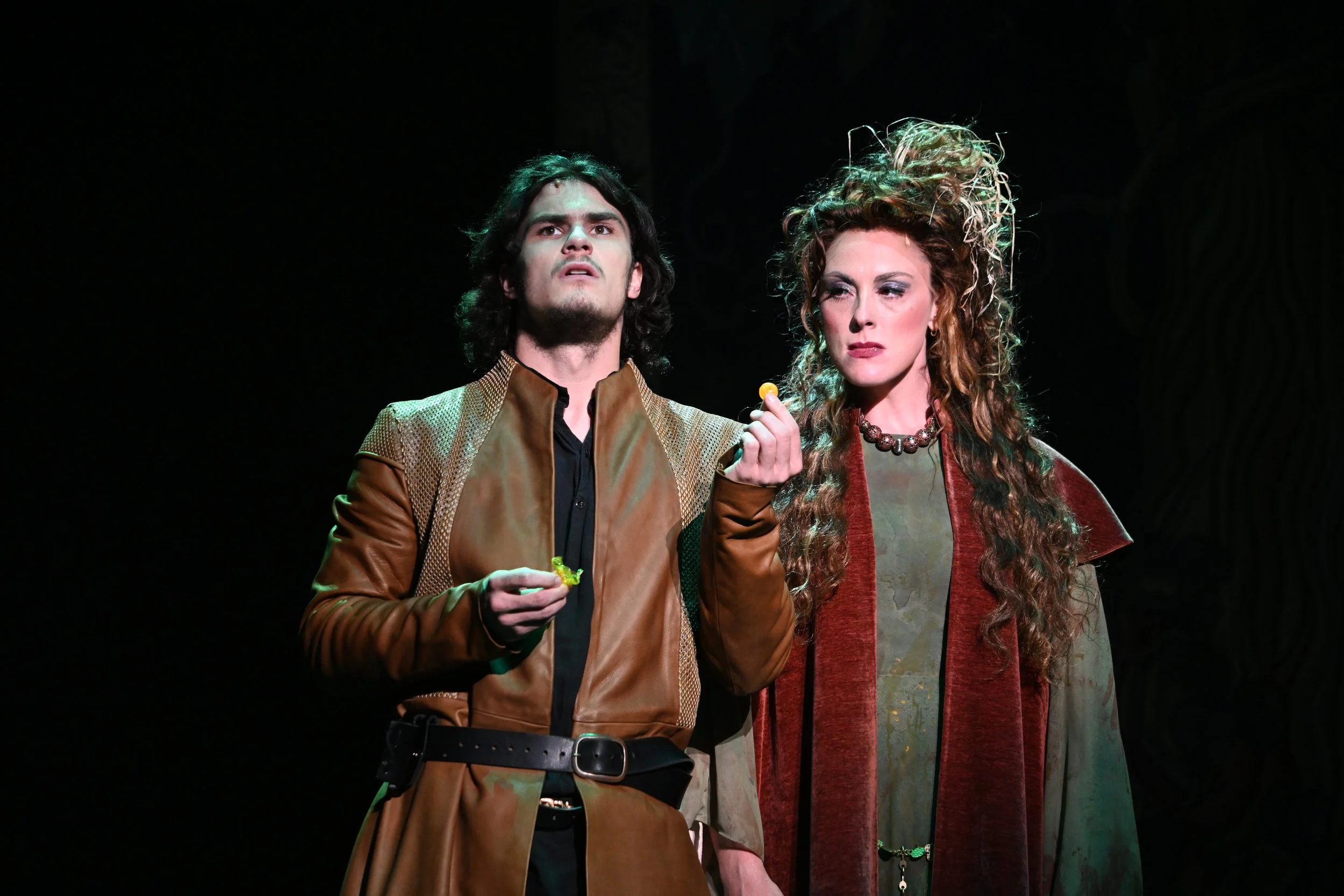 Two actors in costume perform on stage, one man with long dark hair and a brown leather jacket, and one woman with curly hair and a long, multicolored dress, standing in front of a dark background.