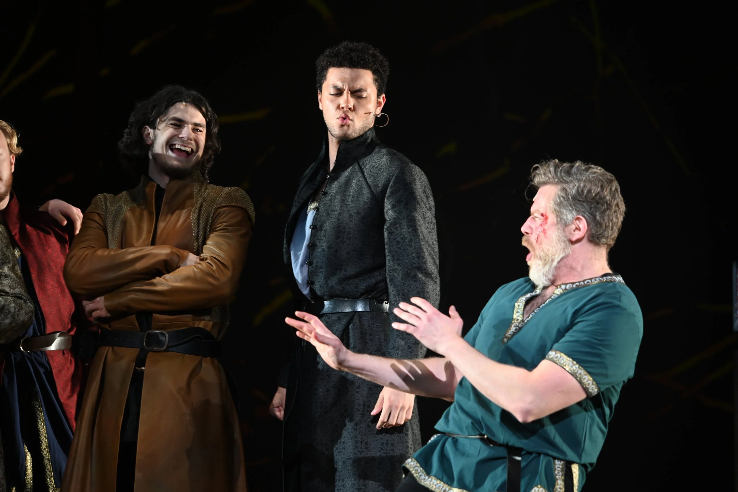 Four men in costumes on stage, with three of them laughing and one gesturing with his hands, during a theatrical performance.