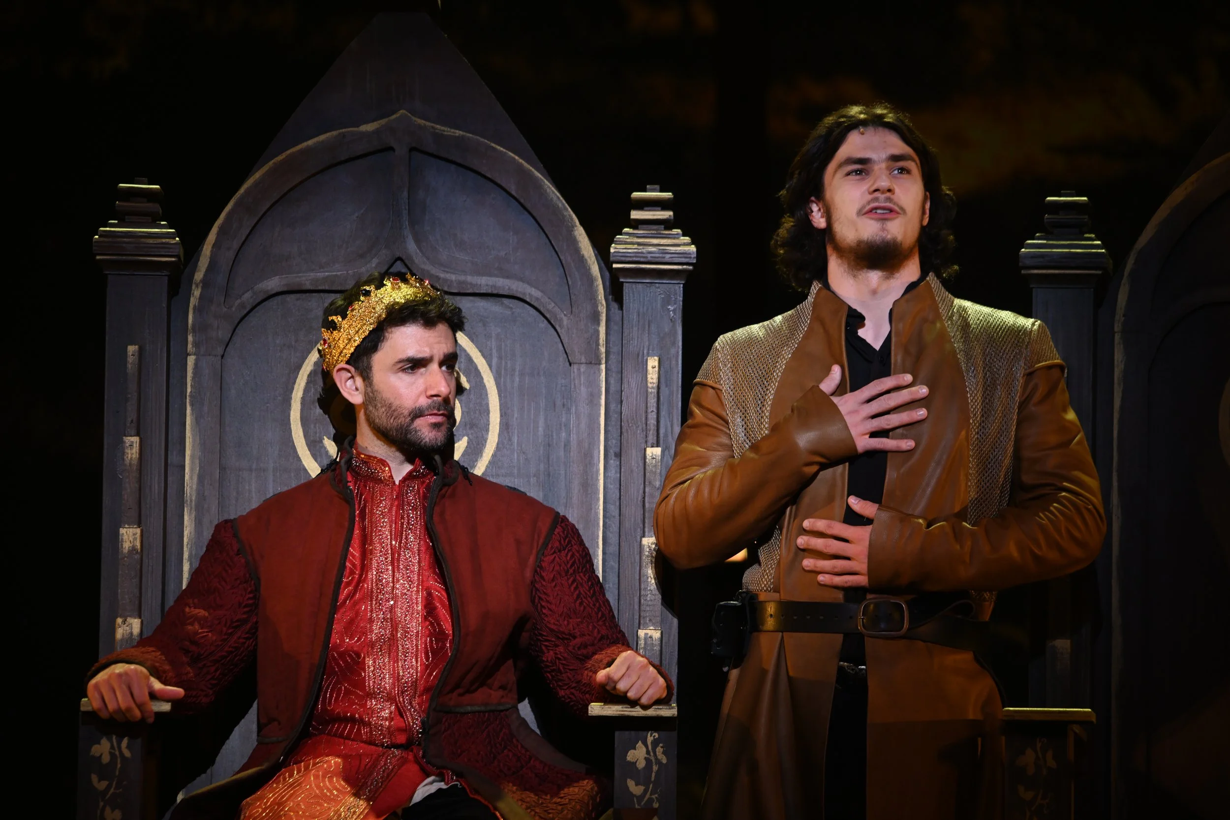 Two men in medieval or fantasy costumes on stage, one sitting on a throne with a golden crown and red robes, the other standing with hand on chest, wearing a brown leather jacket. Dark background.