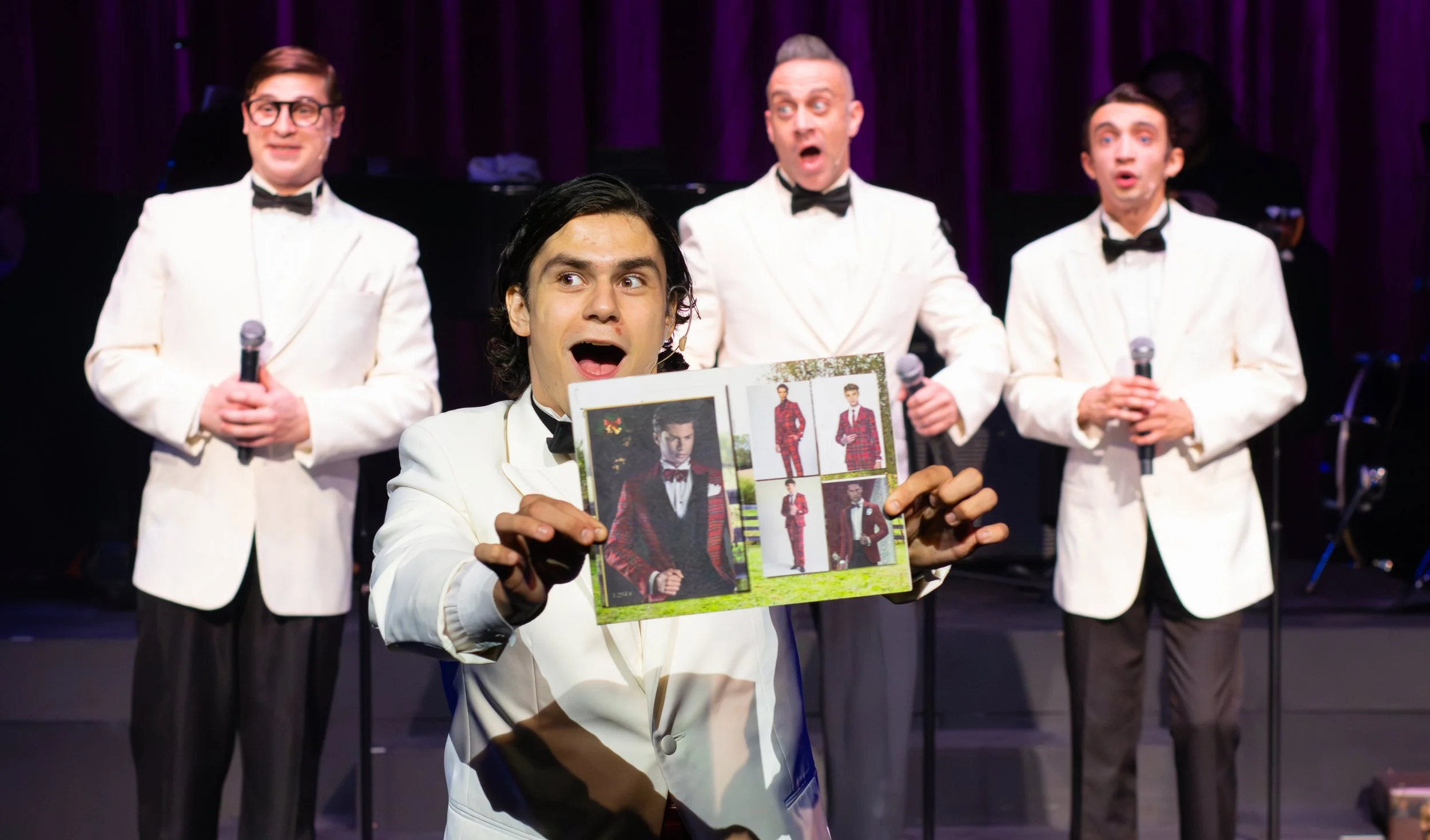 A man in a white tuxedo holds an open photo album showing photos of a man in a red plaid suit, in front of four men dressed in white tuxedos with black bow ties, singing on stage with a dark purple curtain background.