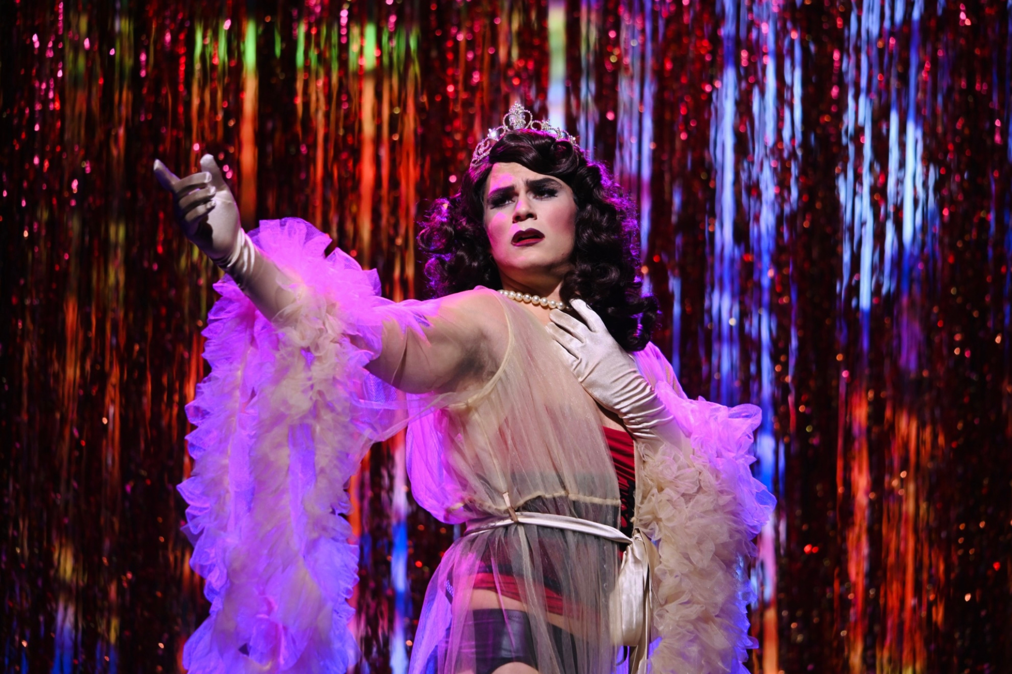 A drag queen performing on stage, wearing a sheer pink robe with ruffled sleeves, a red and black striped outfit underneath, a pearl necklace, a tiara, and white gloves. They are striking a dramatic pose with one arm outstretched and the other hand o