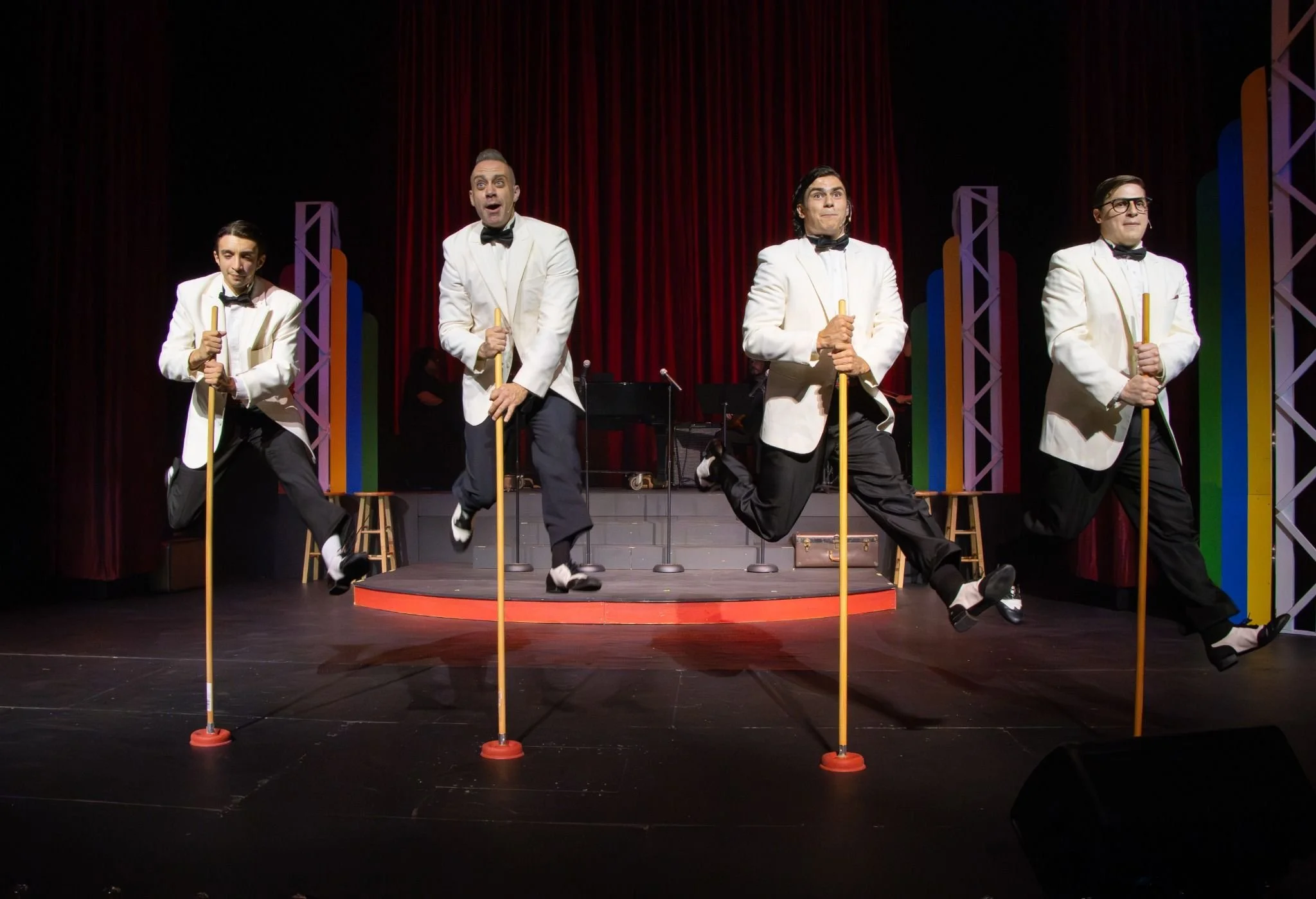 Four male performers in white tuxedos with black bow ties performing a synchronized dance move on stage, holding yellow canes, with a red curtain backdrop and colorful vertical decorations behind them.