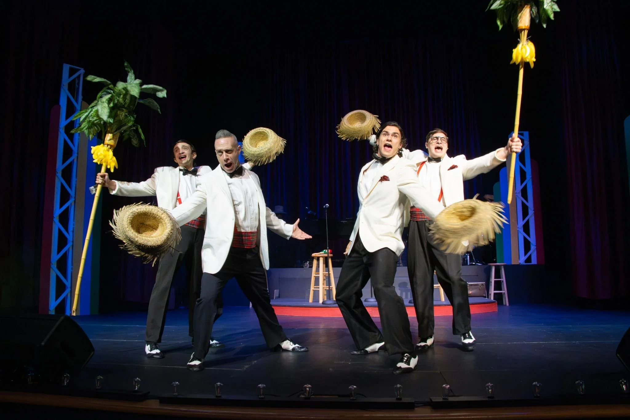 Five men dressed in white tuxedos and black pants performing a tap dance on stage, holding large straw hats and palm trees with yellow flowers.