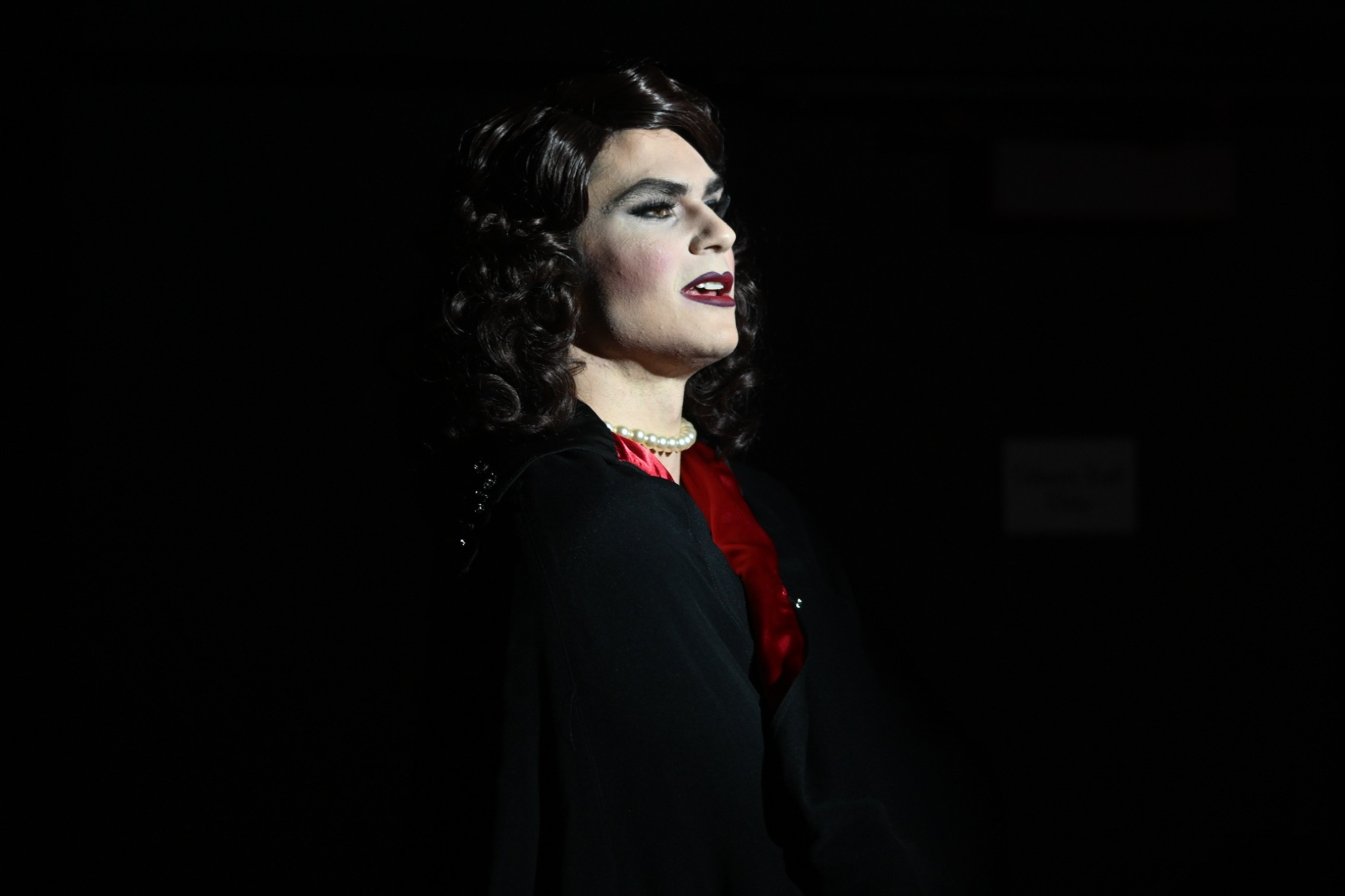 A person in drag with dark, wavy hair, wearing a black outfit with a red inner shirt, pearl necklace, and red lipstick, standing in a dark environment.