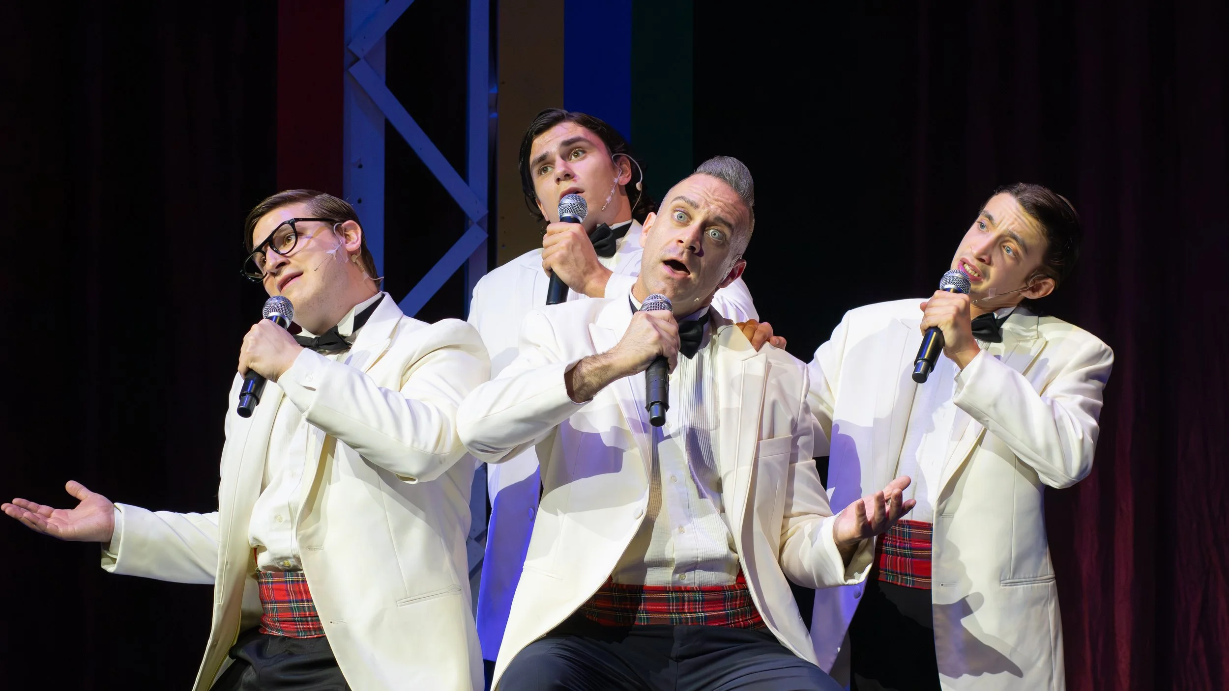 Four men dressed as a musical group called The Four Seasons, wearing white tuxedos with plaid red and black waistbands, performing on stage with microphones, expressive singing and acting.