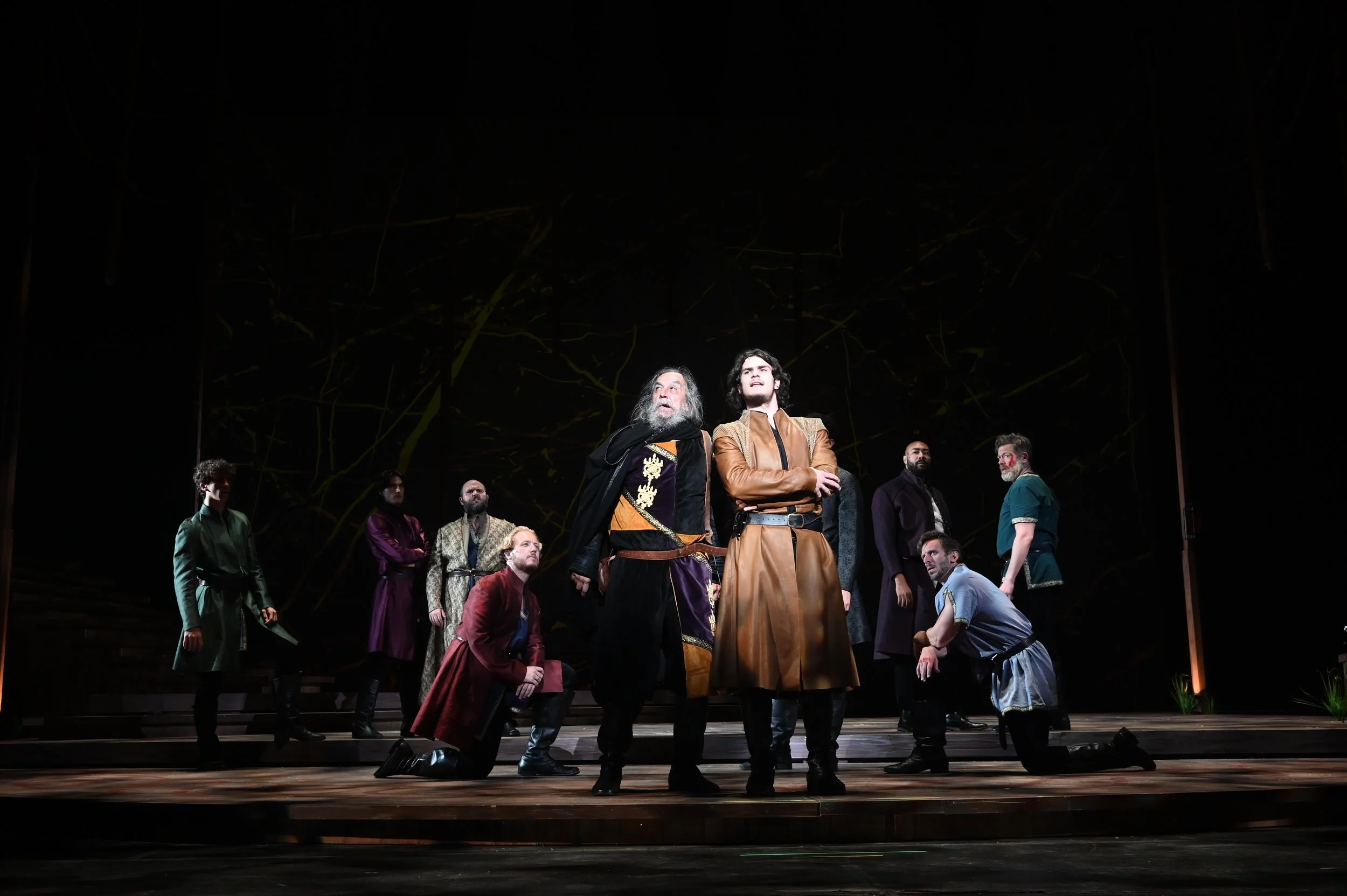 A theatrical stage with nine actors in historical costumes, with two individuals in the foreground and seven behind them, under a dark backdrop with abstract yellow-green lines.