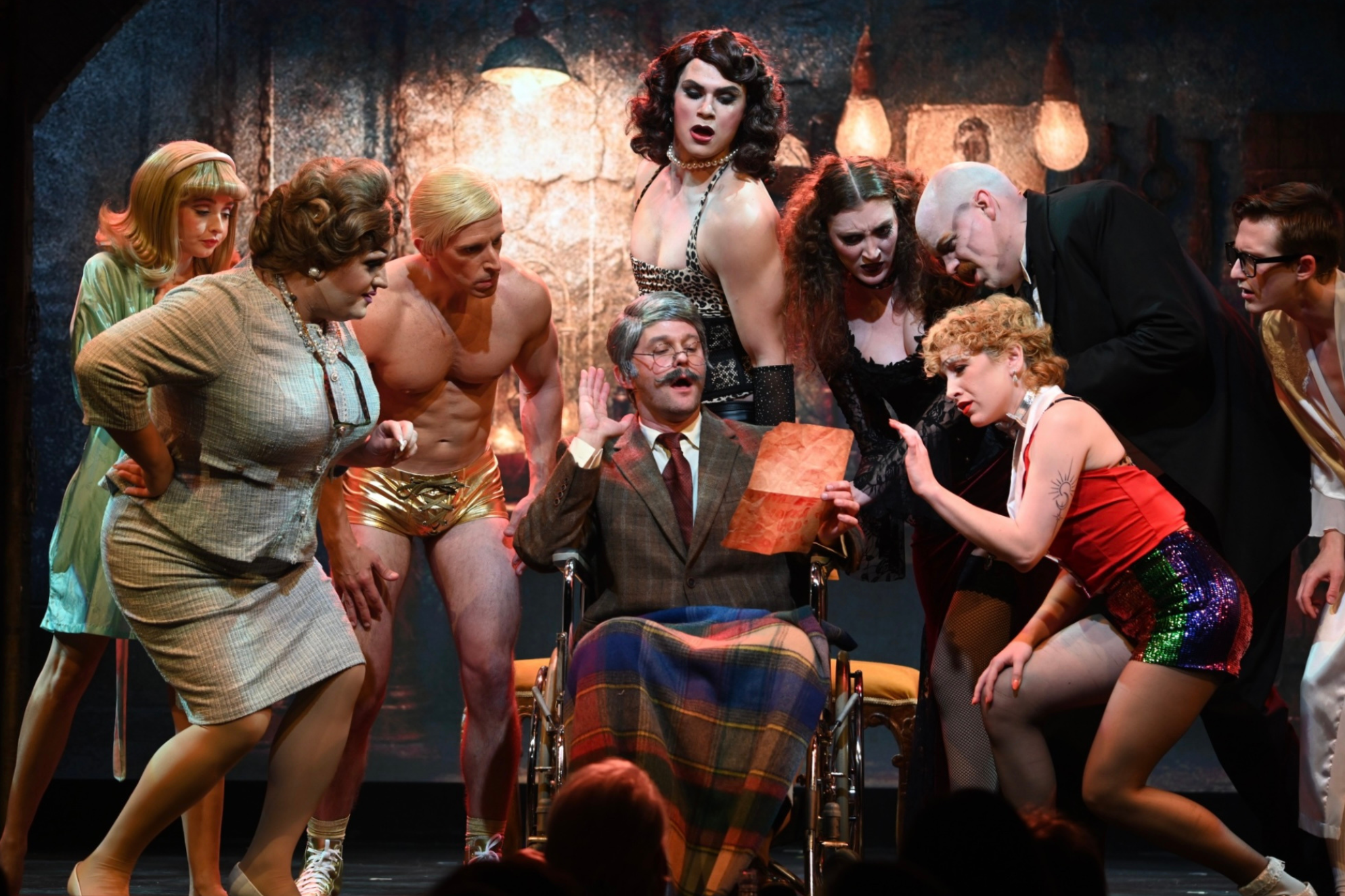 Stage performance with a man in a wheelchair reading a paper surrounded by multiple actors in vintage and eccentric costumes, with expressive gestures and dramatic expressions.