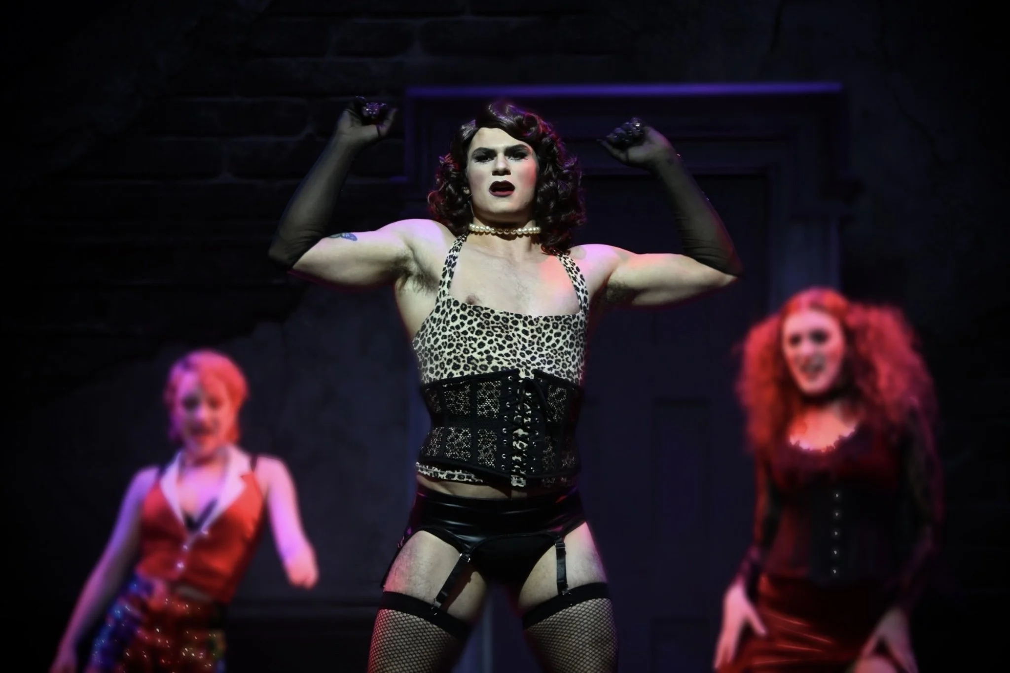 A man dressed in leopard print top, black garter belt, stockings, and gloves performs a show, flexing his muscles on stage with two women in the background.