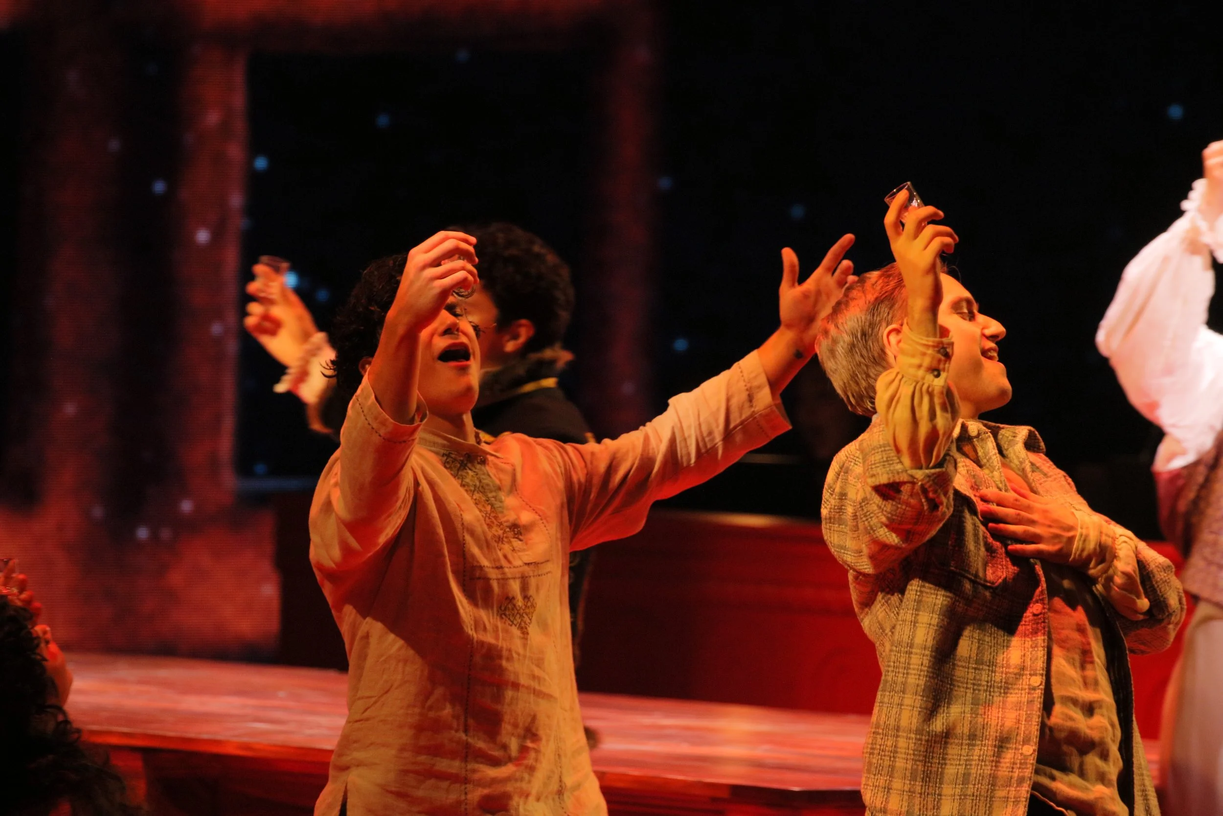 Two women performing on stage, one with glasses and curly hair, singing with eyes closed and arms raised, the other with short gray hair, smiling and holding her hand to her chest. Stage lighting is warm, with a dark background.