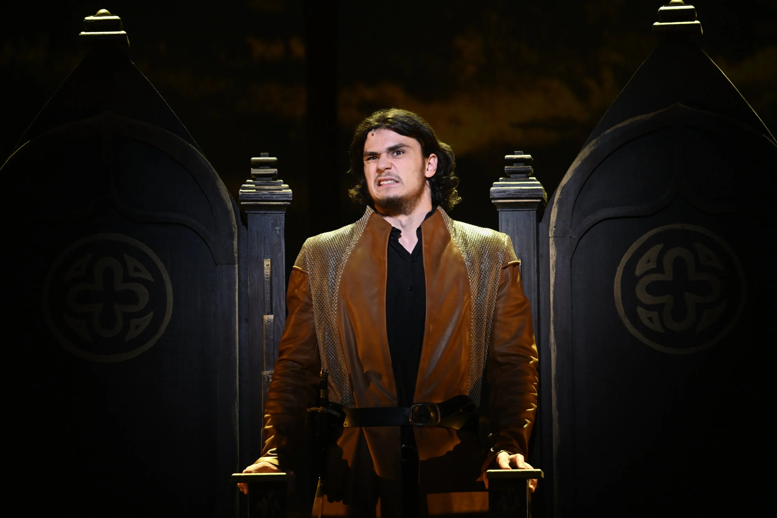 A young man with dark, curly hair stands with an intense, angry expression on stage, in front of a dark gothic black throne with intricate designs, wearing a brown leather and gold-colored jacket.