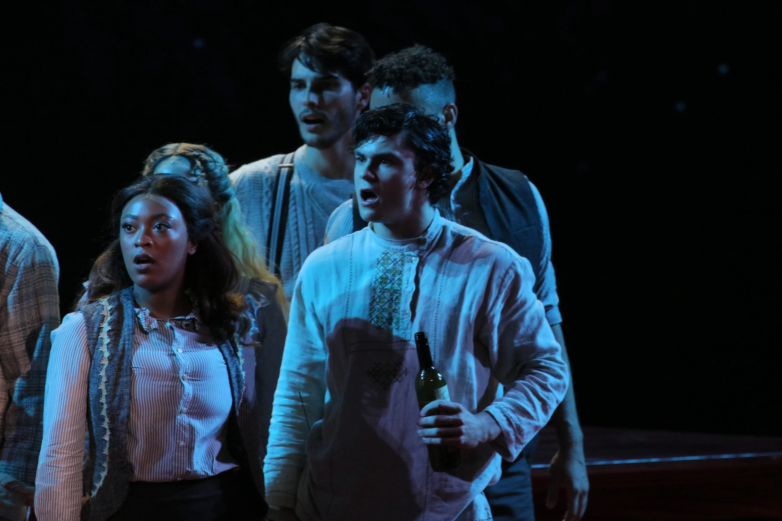 Group of actors on stage in costume, appearing surprised or shocked during a theatrical performance, with a dark background.