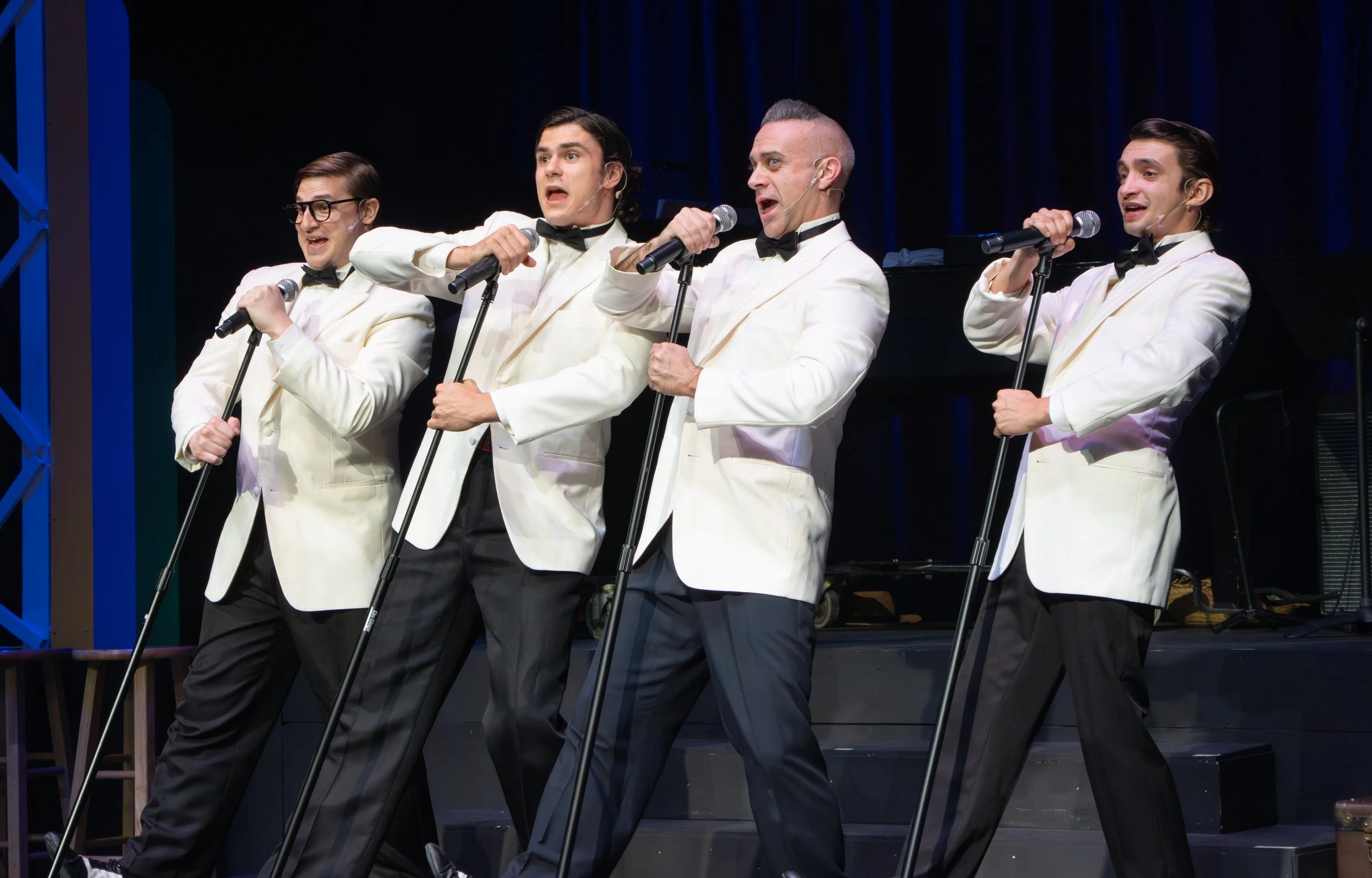 Four male performers on stage dressed in white tuxedos with black bow ties, singing into microphones, with a dark background and stage equipment visible.