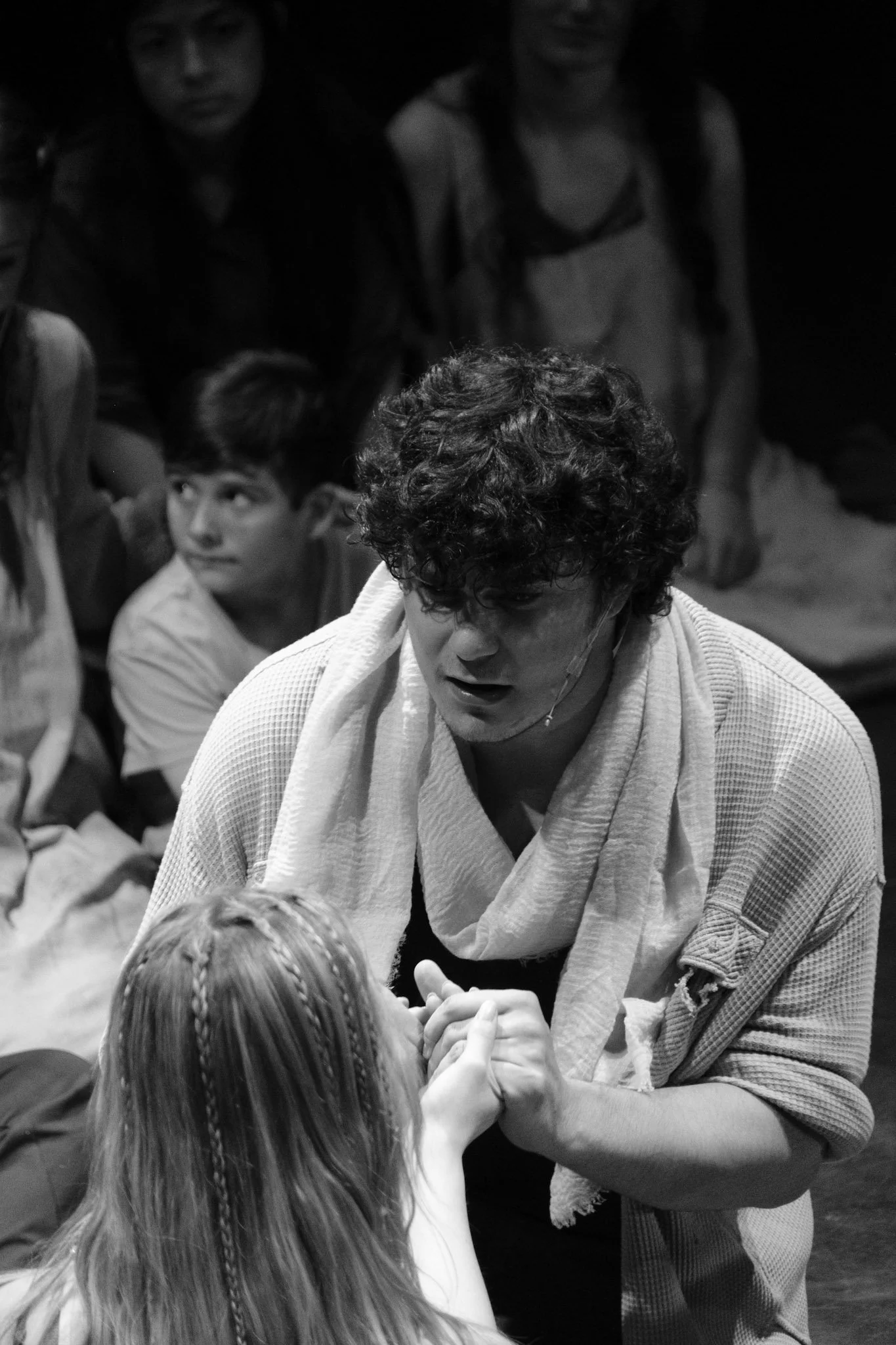 A man with curly hair and a towel around his neck is leaning forward and holding hands with a young girl with braided hair. Several children are seated around them, and they appear to be engaged in a dramatic or emotional scene.